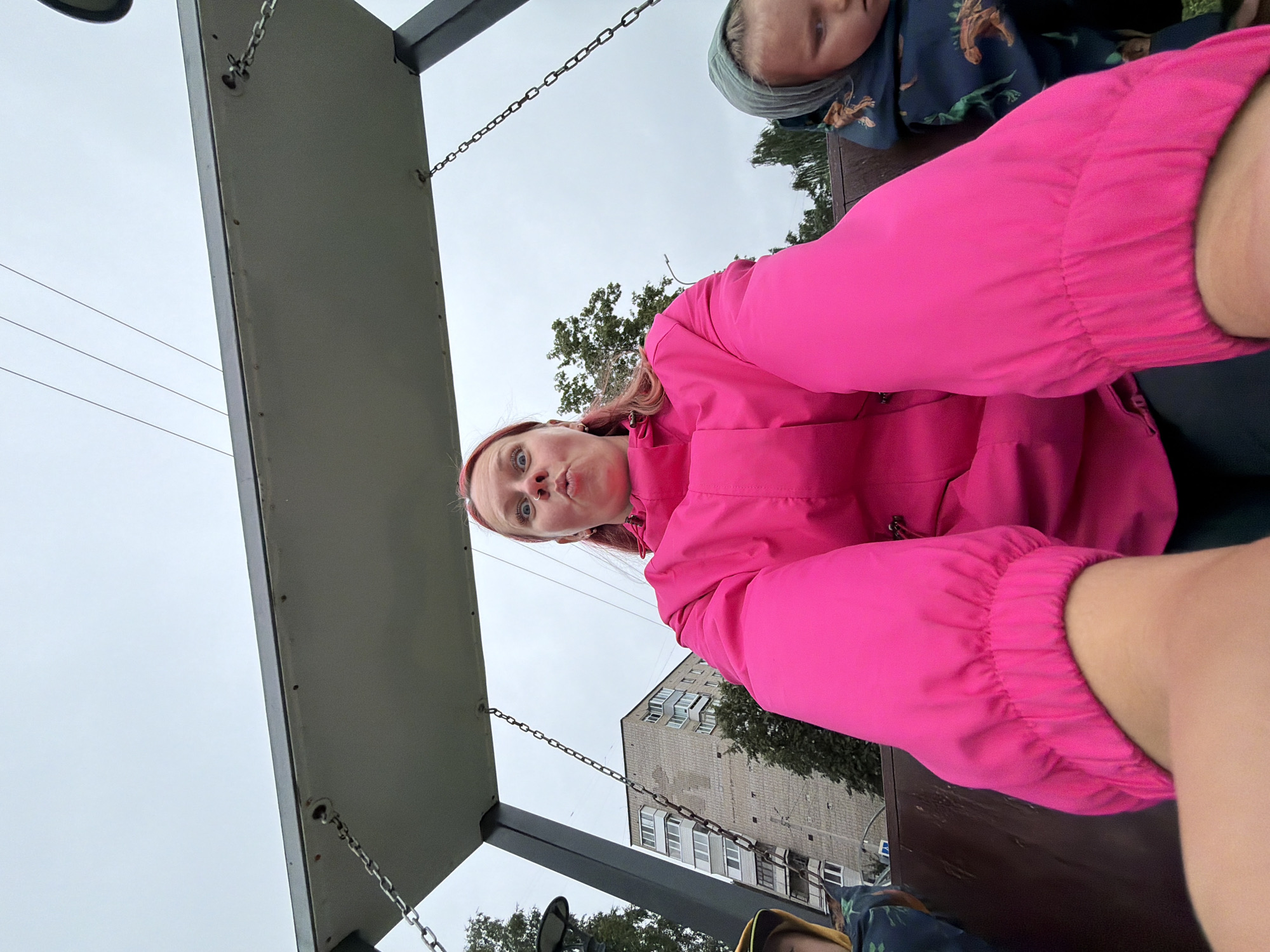 A first-person perspective selfie captures a woman with bright pink hair, wearing a vibrant pink jacket, making a fish-face expression while seated on a swing. To her left, a child in a yellow hat and dark patterned jacket is partially visible. To her right, another child, facing away from the camera, wears a gray hood and a dark jacket with a dinosaur print. The swing is suspended from a metal frame, and the background reveals a cloudy sky, trees, a street lamp, and a multi-story brick apartment building, characteristic of Chaikovsky, Russia. The building's facade shows several balconies and windows. The overall scene suggests an outdoor recreational area.