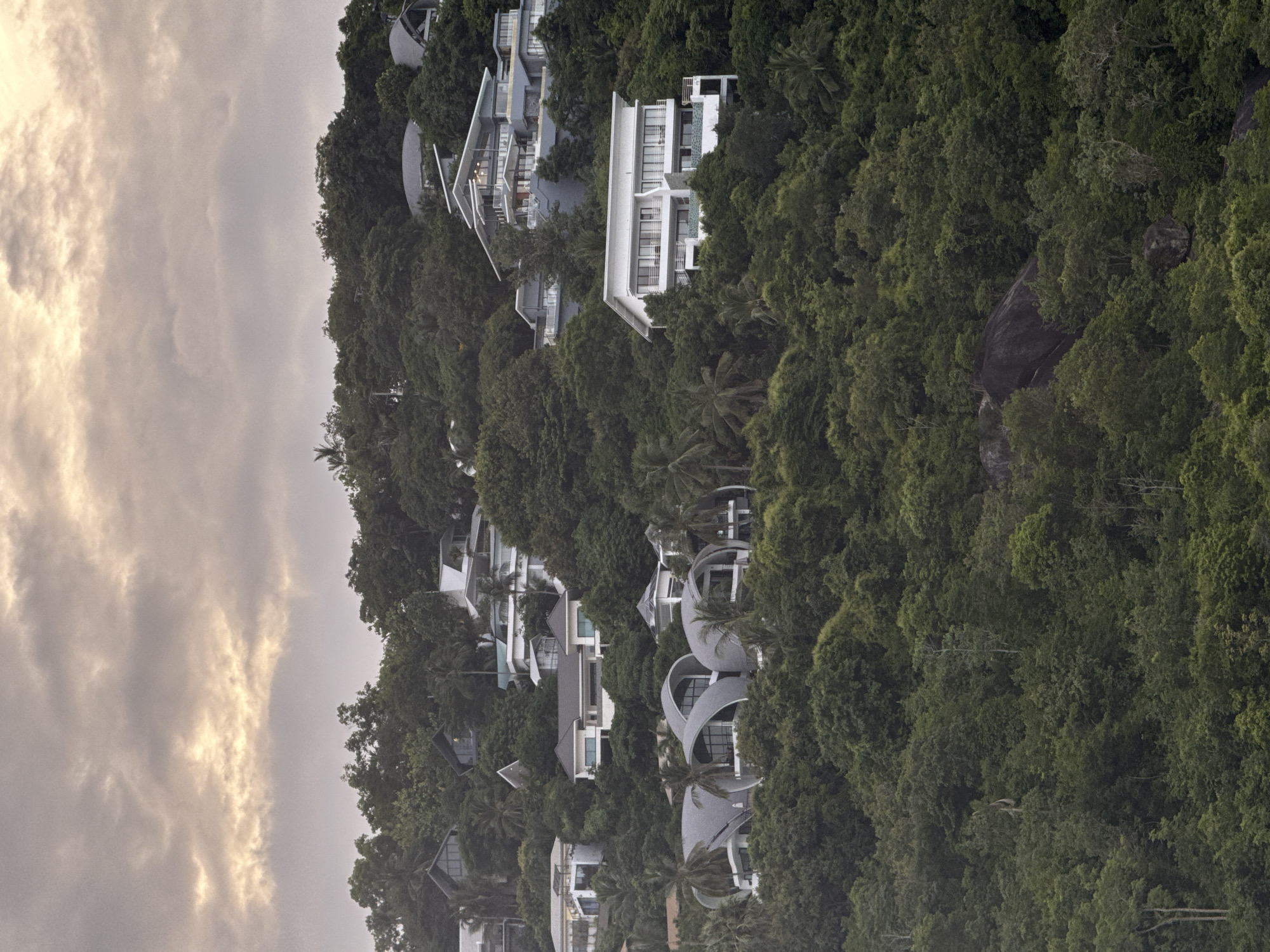 A long shot captures a cluster of modern, luxurious villas nestled into a dense, green hillside on the island of Ko Samui, Thailand. The sky above is overcast with dramatic clouds tinged with the warm glow of sunset. The villas feature distinct architectural styles, from angular, multi-tiered structures with balconies and large windows to more curved, organic designs with gray, shell-like roofs. The lush tropical foliage of the island surrounds and partially obscures the buildings, with palm trees interspersed amongst the dense green canopy. The overall impression is one of opulent tropical living integrated with the natural landscape.