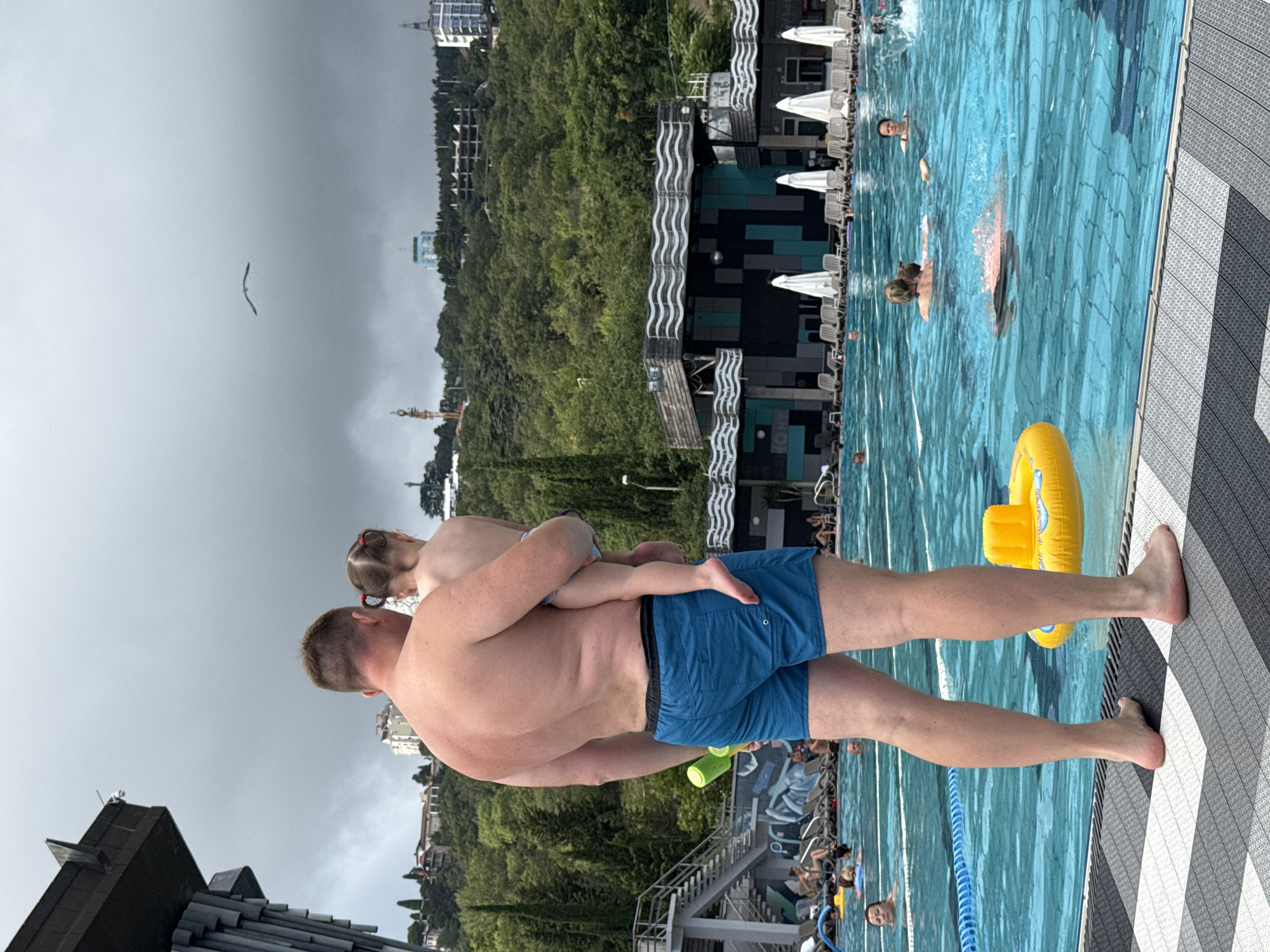 In the city of Yalta, Ukraine, a man holding a young child stands by a large outdoor swimming pool. The man is wearing blue swimming trunks and the child, facing away from the camera, is being held against his chest. The pool is filled with people swimming and relaxing, with a yellow inflatable ring floating in the foreground. In the background, a densely wooded hillside rises, dotted with buildings and structures, and a dramatic, overcast sky looms overhead. A single bird is visible in flight against the cloudy sky.