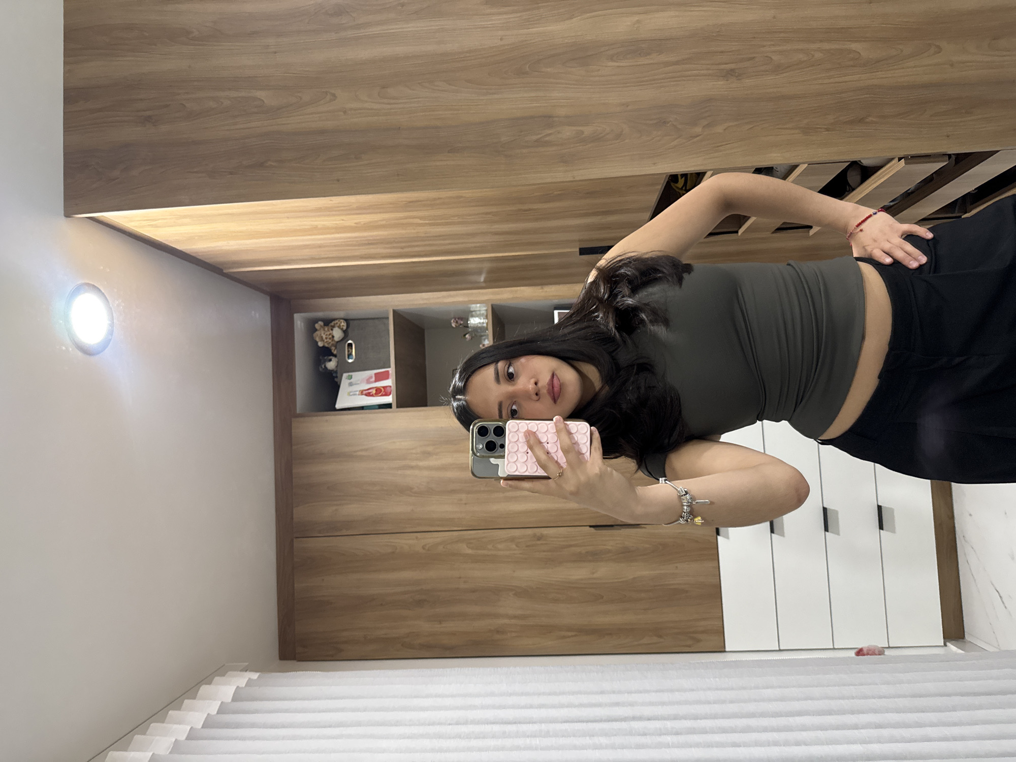 An individual is depicted taking a mirror selfie indoors. The subject is a woman with long dark hair, wearing a dark olive-green sleeveless crop top and black wide-leg pants. Her right hand holds an iPhone with a pink, textured pop-it style case, positioned to capture her reflection. A multi-charm bracelet is visible on her right wrist, and a red string bracelet with a blue bead is on her left wrist. Her left hand rests on her hip.
The background consists of a modern interior space, likely a dressing room or walk-in closet. White walls and ceiling are visible, with a single recessed circular light fixture illuminating the scene. To the left, a white vertical blind or curtain hangs. The majority of the background is comprised of light brown wood-grained cabinetry, extending from floor to ceiling. Some sections of the cabinetry feature white drawers with black horizontal pull handles. Open shelves within the cabinets display various items: a grey fabric bin containing two stuffed animals (one leopard print, one brown) on an upper left shelf; small glass containers on a lower shelf; and several bottles, some containing red liquid, on an upper right shelf. A pull-out drawer system, partially open on the far right, reveals shoes or other stored items. The floor is covered with white marble-patterned tiles.
The specific location within the city of Puebla, Mexico, cannot be determined from the visual information provided in the image.
