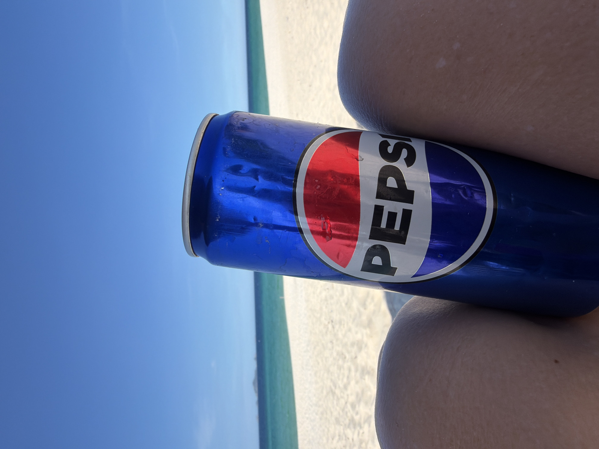 A close-up shot features a Pepsi can held by a person's legs on a sunny beach. The can, partially obscured by the legs, displays the iconic Pepsi logo. In the background, a pristine white sandy beach meets the turquoise ocean under a clear blue sky. In the distance, a small island is visible. The scene suggests a relaxed day at the beach in a tropical or coastal location, possibly Bac Cam Ranh Ward, Vietnam, given the context.