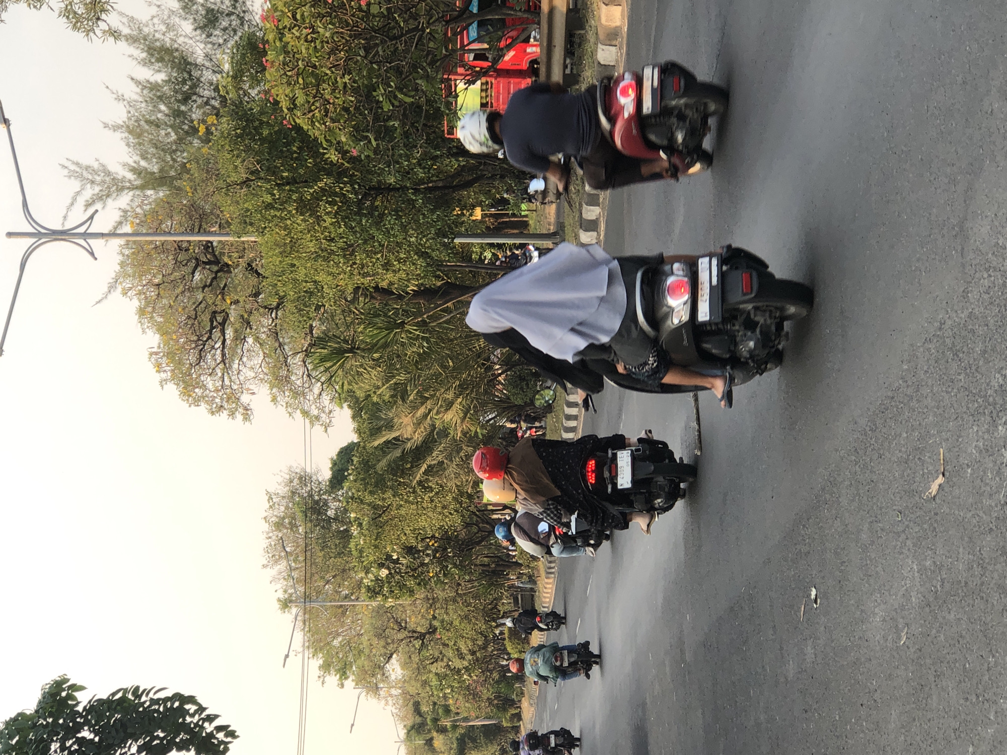 In a sun-drenched urban scene in the city of Ngaban, Indonesia, a procession of motorbikes travels along a paved road lined with lush greenery and trees. The traffic comprises several individuals, predominantly riding scooters or motorcycles, each adorned with helmets, some in vibrant red, blue, or white, others with patterned coverings. The riders are a mix of genders, with a few women wearing hijabs, one in a light blue headscarf and another in a darker covering. One rider in the foreground, a woman in a patterned outfit, is seen from behind, her feet clad in flip-flops resting on the scooter. Another individual on a red scooter, also wearing a white helmet, is visible in the right side of the frame. Further down the road, other motorbikes and their riders are depicted, with one scooter displaying a license plate that reads "N 4309 TEV." The environment suggests a warm climate, with the sky a pale, hazy white. Tall streetlights with a distinctive curved design are interspersed among the trees, and a red utility truck is partially visible on the right side of the image. The road surface shows signs of wear and tear, with scattered leaves and debris.