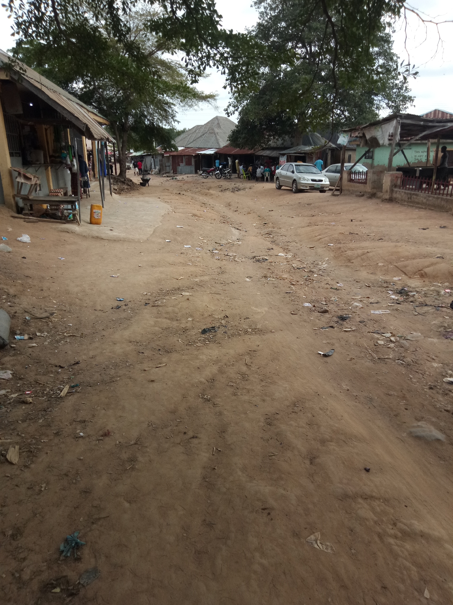 The image depicts a dirt road in what appears to be a semi-urban or rural area. On the left side of the road is a low-lying structure with a corrugated metal roof, resembling a shop or market stall. Several people are visible near the stall, and some items, including what looks like eggs in a carton, are displayed on a wooden table outside the structure. A yellow plastic container is also present. Further down the road, a silver sedan car is parked, and another vehicle, possibly a motorcycle, is visible in the distance. The road itself is unpaved and shows signs of wear and tear, with scattered debris, including plastic bags, paper, and other refuse, along its surface. The environment is characterized by dusty soil, sparse vegetation, and a cloudy sky. Buildings with metal roofing are visible on both sides of the road, suggesting a settlement or village. The overall impression is one of a functional, albeit unkempt, community pathway.