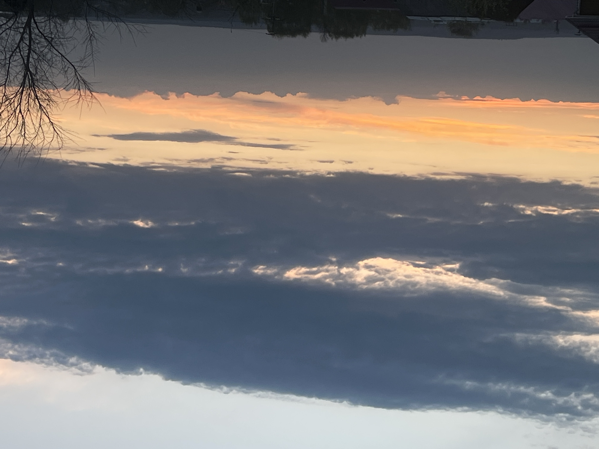 The image depicts a sunset scene, possibly in Votkinsk, Russia, based on the information provided. The photo is oriented upside down, with a body of water and cityscape visible at the top, suggesting the photo was taken near a lake or river. Predominantly, the view is of the sky, filled with layers of clouds. The lower portion of the sky shows dark, heavy cloud formations. Above these, a band of orange and yellow hues indicates the setting sun, casting light onto the clouds. A bare tree branch appears silhouetted in the upper-left corner. The overall mood is tranquil, highlighting a moment of natural beauty.
