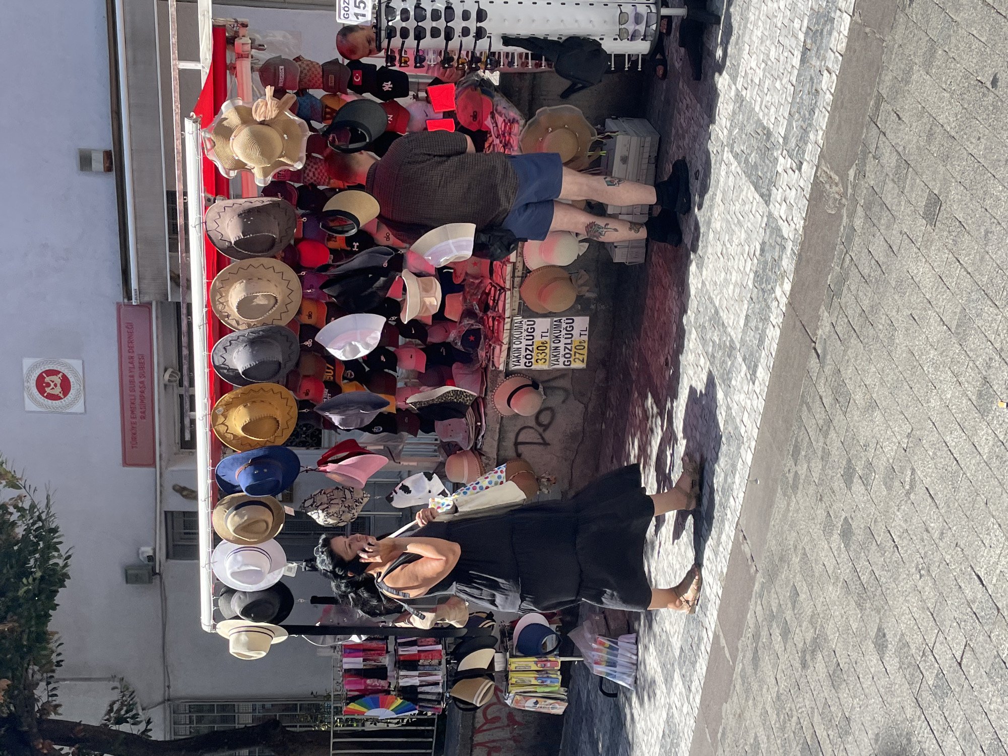 A street vendor stall is set up on a cobblestone sidewalk in Kadıköy, Turkey, displaying a variety of hats and sunglasses. A woman in a dark, tiered dress is walking past the stall while talking on her phone. A man with tattoos on his legs is standing near the stall, looking at the merchandise. The stall is covered by a red awning and features shelves and hanging displays packed with hats of different styles and colors, including baseball caps, sun hats, and fedoras. A rack of sunglasses is also visible on the right side of the stall. The background shows a building with a sign that appears to be in Turkish. The scene is lit by bright daylight, casting shadows on the ground.