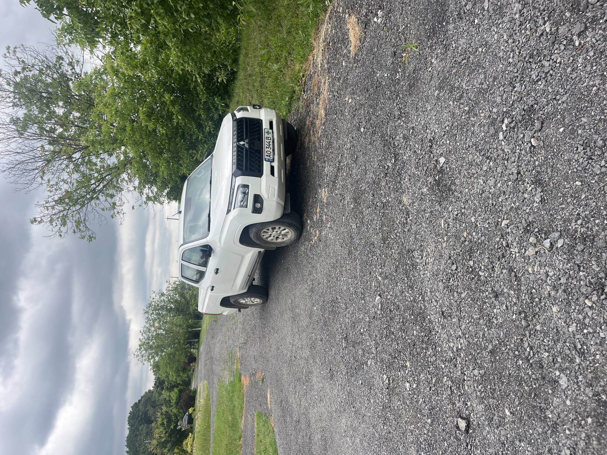 A horizontally oriented outdoor photograph depicts a white Mitsubishi pickup truck parked on a gravel surface, flanked by green vegetation and trees under an overcast sky. The truck is positioned facing the viewer's left, with its front visible in the mid-ground.
The primary subject is a white Mitsubishi pickup truck, clearly identifiable by the "MITSUBISHI" text visible on its front grille. A Ukrainian license plate, "AO 344 B," featuring the national flag, is affixed to the front bumper, indicating the setting is in Ukraine, consistent with the provided location context of Onokivtsi. The truck has light-colored multi-spoke wheels and appears to be a double-cab model.
The setting is a rural or semi-rural area, characterized by a wide gravel road or driveway occupying the foreground and right side of the image. Patches of green grass are interspersed along the edge of the gravel. Lush green trees form a dense backdrop, particularly on the left side of the frame and in the distance. The sky is largely cloudy, suggesting an overcast day. No human activity is visible; the truck appears to be parked. The overall impression is one of a quiet, natural environment.