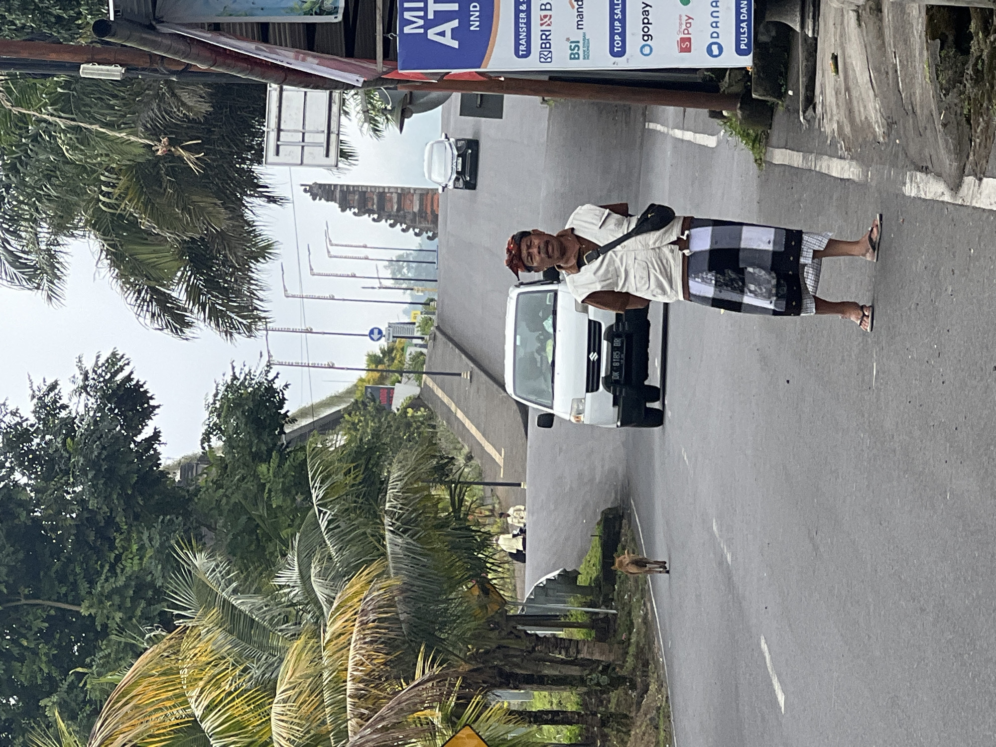 A man stands on the side of a road in Rendang, Indonesia, wearing traditional Balinese clothing. He has a red and gold patterned headscarf, a light-colored vest over a brown shirt, and a black sarong. He wears dark flip-flops and has a black bag slung across his chest. Behind him, a white van with a license plate starting with "DK" is parked on the road. Further up the road, a dark-colored car is in motion. The road is lined with palm trees and dense green foliage. In the background, a traditional Balinese temple gate stands tall. The sky is overcast. A brown dog walks away from the camera on the left side of the road. A sign on the right side of the road displays "MINI ATM" and various financial service logos. The scene appears to be a typical street view in a tropical region.