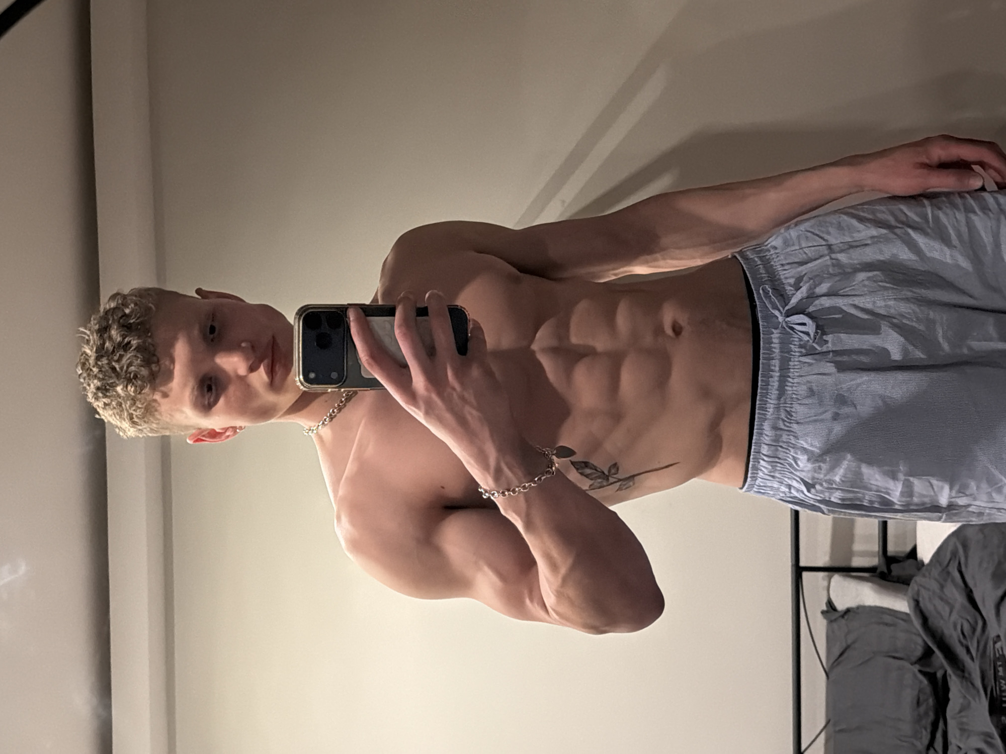 This media file is a mirror selfie featuring a young man indoors. The subject has light, curly hair, fair skin, and a highly muscular physique with visible abdominal definition and prominent veins in his arms. He is shirtless, wearing light blue, pinstriped shorts or pajama bottoms. He accessorizes with a silver chain necklace and a matching silver bracelet on his right wrist. A tattoo depicting a stylized branch or leaf design is visible on his left side, below the rib cage.
The man is holding a dark-colored smartphone in his right hand, angled to capture his reflection in the mirror. He looks directly into the phone's camera with a neutral expression. The background primarily consists of a plain, light-colored wall. In the lower right section of the image, some dark, soft objects, possibly clothes or bedding, are partially visible, suggesting a bedroom or private living space. The lighting is even and soft, indicating an indoor setting potentially lit by artificial light or indirect natural light. There is no visible text in the image.