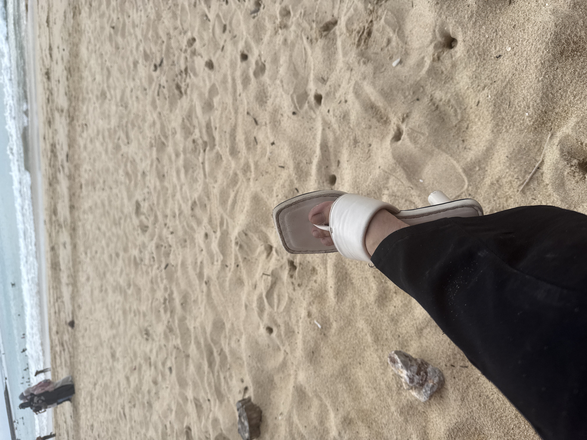 The image shows a person's legs and feet standing on a sandy beach. The person is wearing black pants and white slide sandals. The sand is light brown and has footprints and small pebbles scattered on it. In the background, the ocean water is visible, appearing calm and light blue. The shoreline is wet, suggesting the tide is close. Further in the distance, the waves are breaking gently. The lighting suggests it could be daytime, possibly overcast or early morning/late afternoon given the diffused light. There are no discernible emotions conveyed by the image. The setting is clearly a beach, with the sand and ocean being prominent elements. No text is visible in the image.
