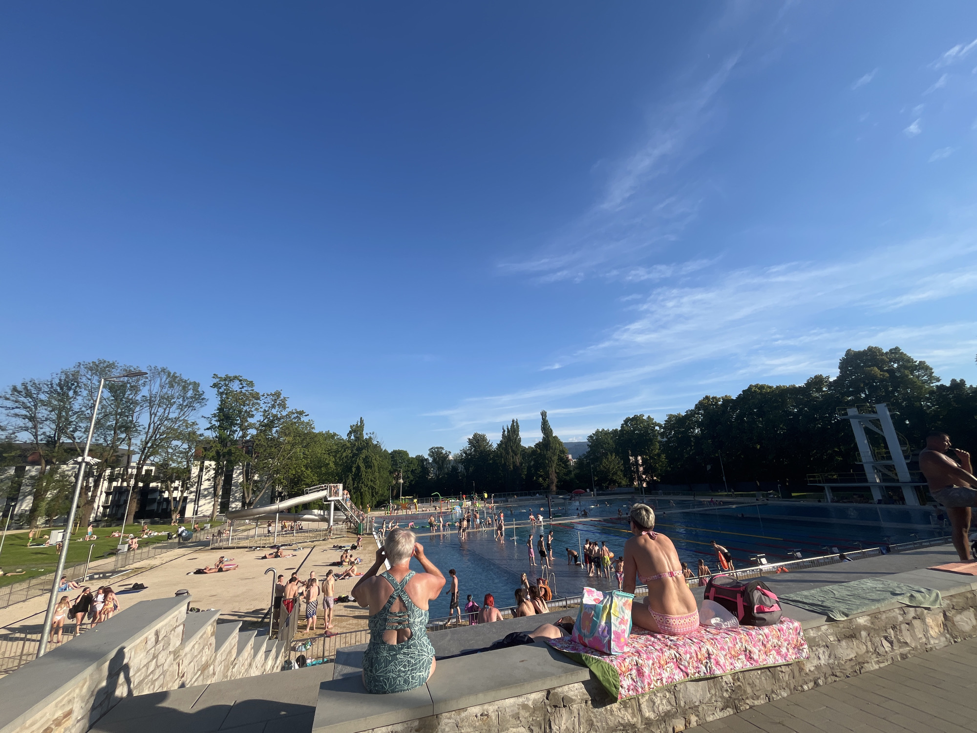 In an outdoor public swimming pool on a sunny day, a man in black swim trunks and a woman in a pink bikini sit facing away from the camera, looking towards the pool. The woman is sitting on a pink and white patterned towel, with a colorful tote bag and a grey backpack beside her. The man is standing. Behind them, the pool is filled with people swimming and wading, with a diving board and a slide visible in the background. Trees and buildings line the perimeter of the pool area.