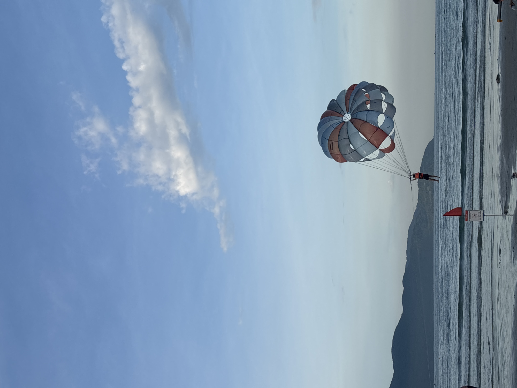 The image depicts a parasailor in mid-air above the ocean, with a colorful parachute catching the breeze. The parasailor, wearing a life vest, is suspended by lines attached to the parachute. The scene appears to be a beach or coastal area, with a calm sea and distant hills or mountains forming the horizon. A red flag is planted in the sand near the water's edge, along with a sign. The sky is partly cloudy with a soft, diffused light, suggesting it might be late afternoon or early morning. The overall impression is one of leisure and outdoor recreation. This scene is likely occurring at one of Đà Nẵng's popular beaches, such as My Khe Beach or Non Nuoc Beach, known for their wide sandy shores and opportunities for water sports.