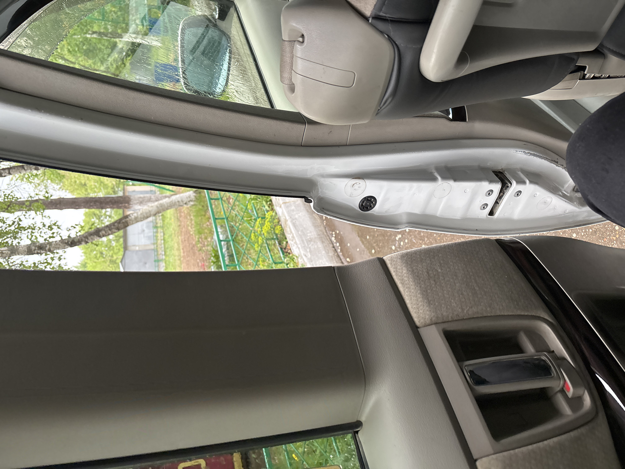 The image shows the interior of a vehicle, specifically the driver's side door frame and a portion of the dashboard. The door is open, revealing the inner workings of the door frame. Visible through the open door is a blurred outdoor scene, including trees, greenery, and a fence. The dashboard area features an air vent. The location is unknown.