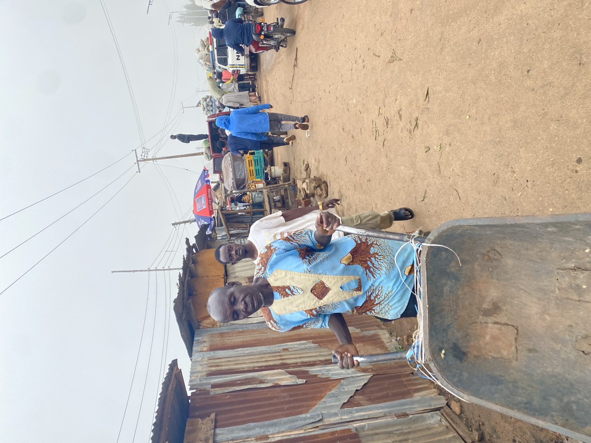The image depicts a bustling outdoor market scene in Gura Top, Nigeria, on an overcast day. In the foreground, an older man with dark skin and a shaved head, wearing a light blue, ornately patterned dashiki, pushes a metal wheelbarrow. He is walking on a dirt path beside a corrugated metal structure. Behind him, a younger man with dark skin, wearing a white t-shirt, walks closely. The path is lined with stalls and various vehicles, including a colorful tricycle-like vehicle, a white police SUV with "NR" on its side, and motorcycles. Several people are seen walking and interacting in the background, some carrying goods. The environment appears somewhat disheveled, with various items scattered on the ground. Power lines crisscross the sky overhead, indicating an urban or semi-urban setting. The overall impression is one of lively commerce and daily activity in a Nigerian marketplace.