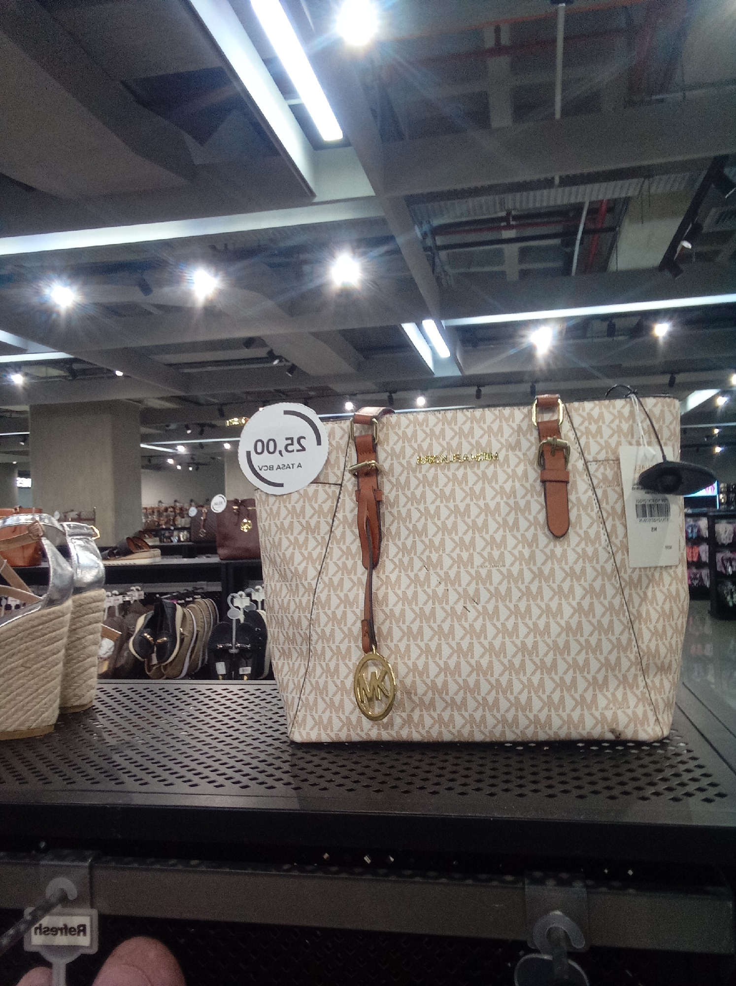 The image shows a beige Michael Kors tote bag with brown leather straps and gold hardware on a black metal shelf. The bag is covered in a repeating "MK" pattern. A price tag is attached to the bag. To the left of the bag, there are silver wedge sandals. Other shoes and bags are visible in the background. The setting appears to be a department store with bright overhead lighting. Based on the image alone, there is no way to definitively confirm this location in the city of Parroquia Guatire, Venezuela.