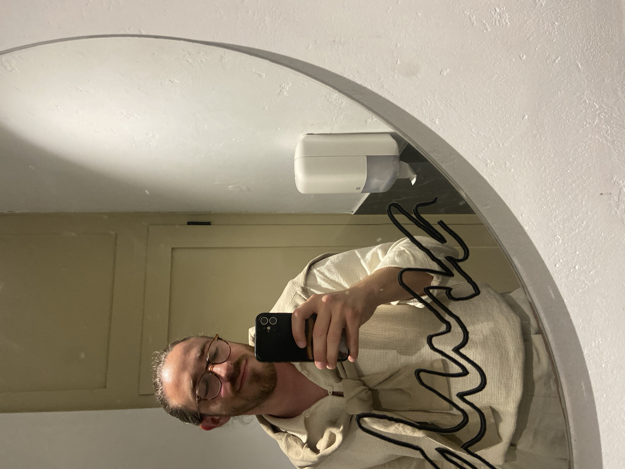 A man is taking a selfie in a mirror in a bathroom. The mirror is oval-shaped and mounted on a white wall. A white soap dispenser is visible above the man's reflection. The man is wearing glasses and a light-colored, textured shirt. He is holding a black smartphone in his right hand. The bathroom walls are a pale color and there appears to be a cabinet or door behind the man's reflection. Location Petrovac centar, Montenegro can not be extracted from image.
