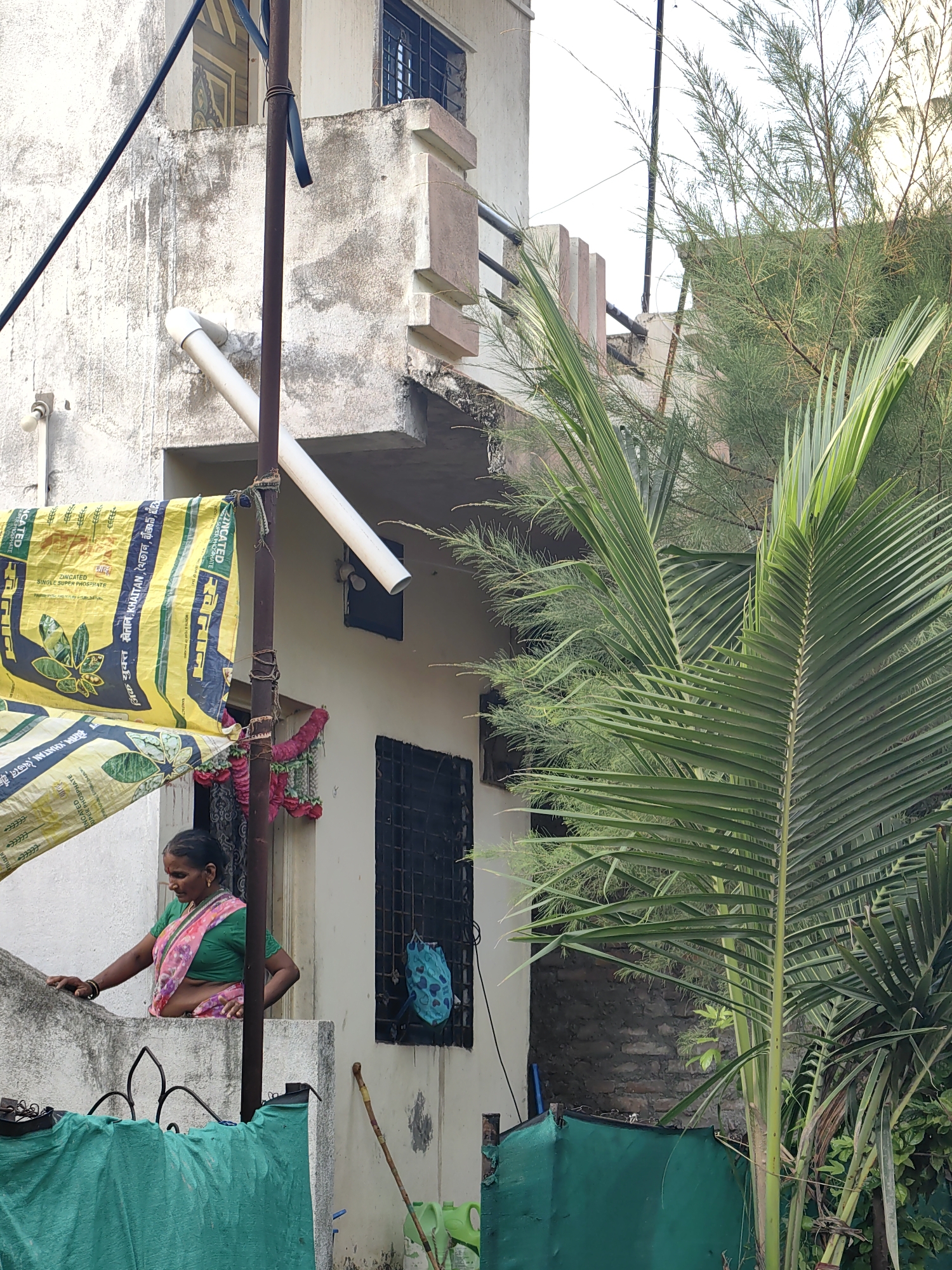 A woman in a green and pink sari stands on a ledge outside a building in Mhaiswadi, India. She is looking down and to her left, with her left hand resting on a low concrete wall. Behind her, a large yellow tarp with writing on it is draped over a pole. The building is made of weathered concrete with a few windows. There are several green plants in the foreground and background, including a large palm frond. A thin metal pole, possibly part of a fence or railing, runs vertically through the center of the image. The sky is overcast.