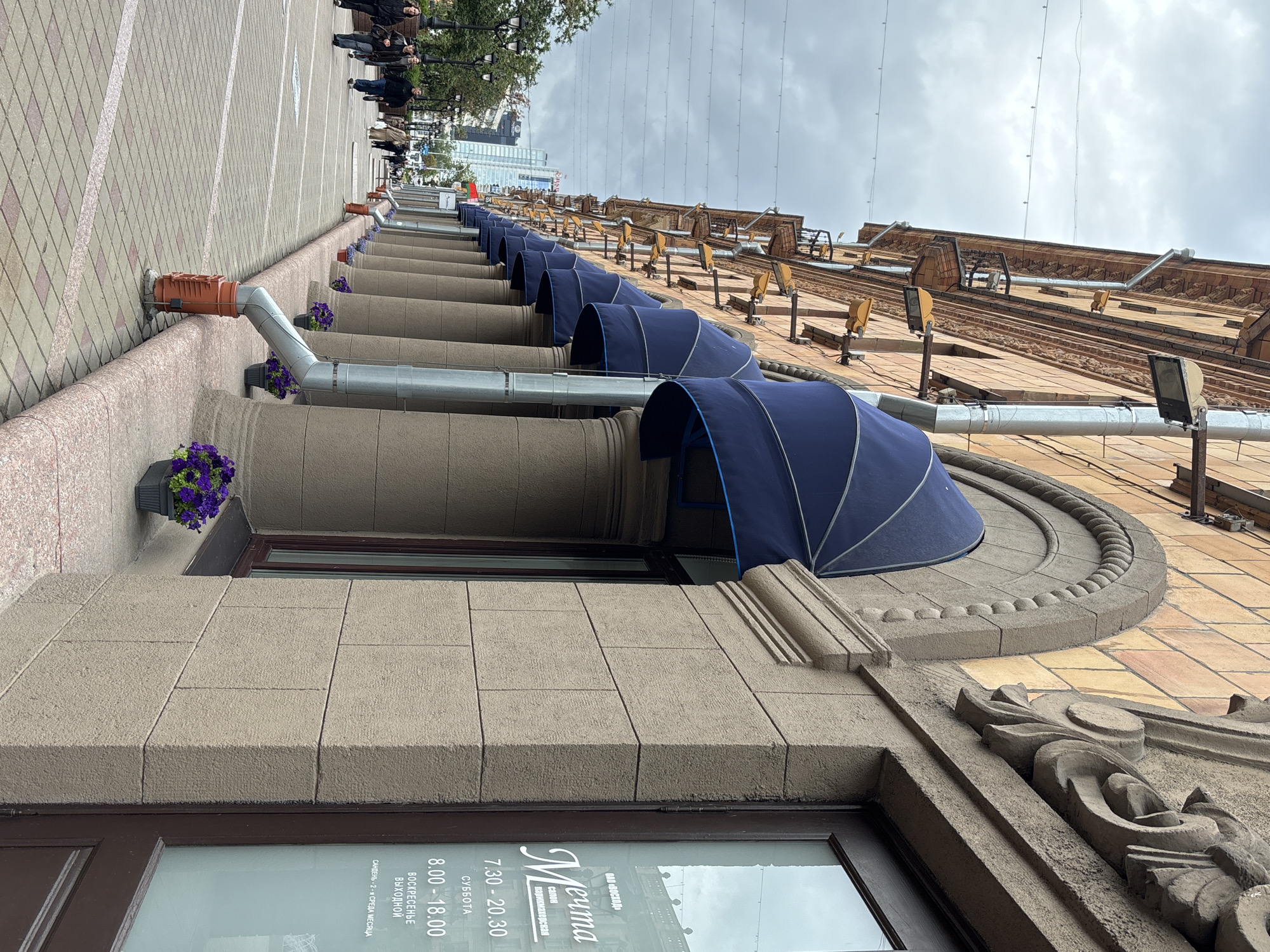 This is a low-angle, eye-level, outdoor, full shot of a street-level storefront with multiple awnings and columns, taken on a cloudy day. The building is made of tan-colored stone blocks and features large, arched windows with blue awnings. A drainpipe runs vertically down the side of the building, supported by an orange plastic fixture at the bottom. The sidewalk is paved with interlocking bricks in shades of pink and grey. Several people are walking on the sidewalk in the background, dressed in casual clothing. In the distance, a modern skyscraper is visible. A sign on the storefront window reads "Мечта салон парикмахерская" and lists operating hours. The overall scene depicts an urban commercial area.
