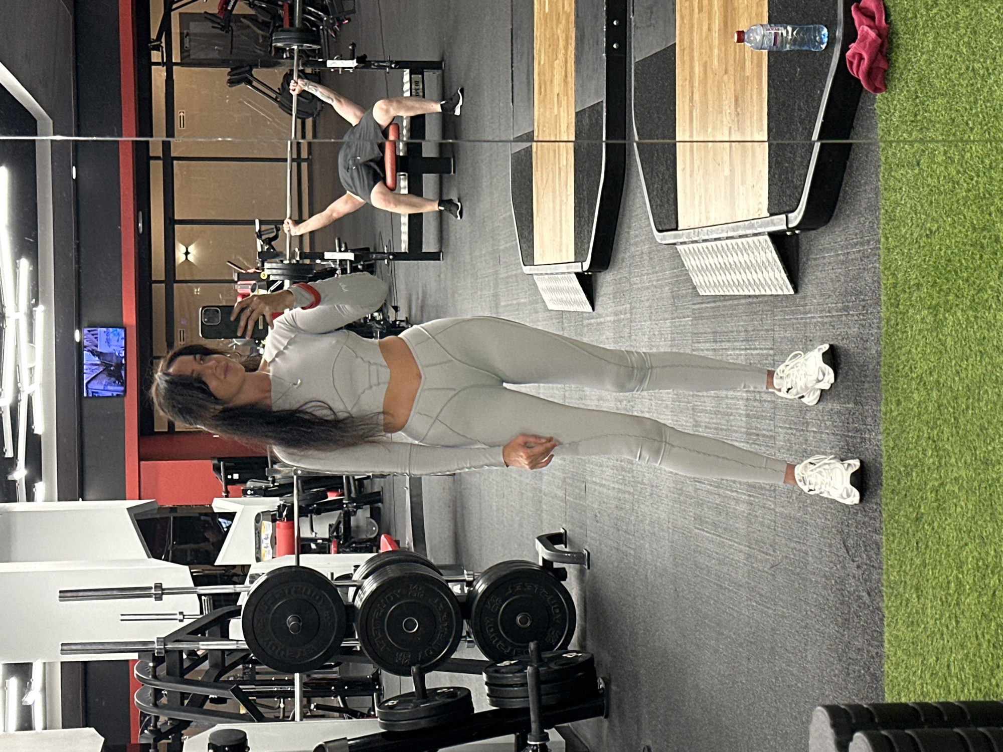 The image depicts a woman in a gym, taking a mirror selfie. She is wearing a light gray athletic outfit, consisting of a long-sleeved crop top and leggings, with white athletic shoes. Her dark hair is down, and she is holding a smartphone in her right hand. In the background, reflected in a large mirror, a man is seen performing a bench press. The gym environment includes weight racks with plates, exercise equipment, and a black floor with areas of artificial turf. The overall lighting suggests an indoor setting, typical of a fitness facility. There is no information in the image to suggest this location is in Irkutsk, Russia.