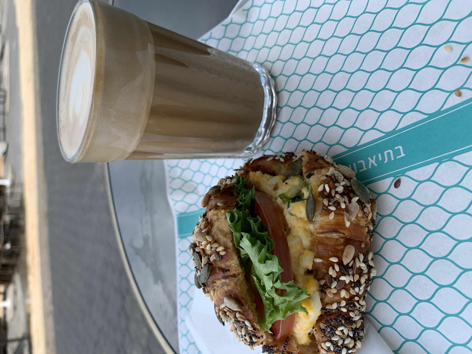 An elevated, close-up view depicts a breakfast or lunch setting at an outdoor café in Tel-Aviv, Israel. The primary subjects are a savory croissant sandwich and a glass of latte, positioned on a dark, round table.
The croissant, split horizontally, is generously coated with a mix of poppy seeds, sesame seeds, sunflower seeds, and pumpkin seeds. Its filling consists of a visible layer of egg salad with green flecks (likely herbs), topped by two slices of red tomato, and finished with a fresh green lettuce leaf.
Adjacent to the croissant is a tall, clear glass containing a layered latte. The beverage displays a light brown liquid base with a thick layer of frothed milk on top, featuring faint latte art resembling a heart.
Both items rest on a white paper liner with a repeating teal chain-link pattern. A solid teal stripe runs diagonally across the paper's bottom right, bearing the white Hebrew text "בתיאבון" (B'Teavon), meaning "Bon Appétit."
The table surface is dark gray or black, possibly glass or polished metal, reflecting light. The blurred background indicates an outdoor urban environment, with a dark gray paved ground directly beyond the table, followed by a lighter-colored path or area. Faintly visible blurred structures suggest outdoor furniture or building elements, consistent with a street-side or patio café setting. No people, actions, or specific interactions are visible.