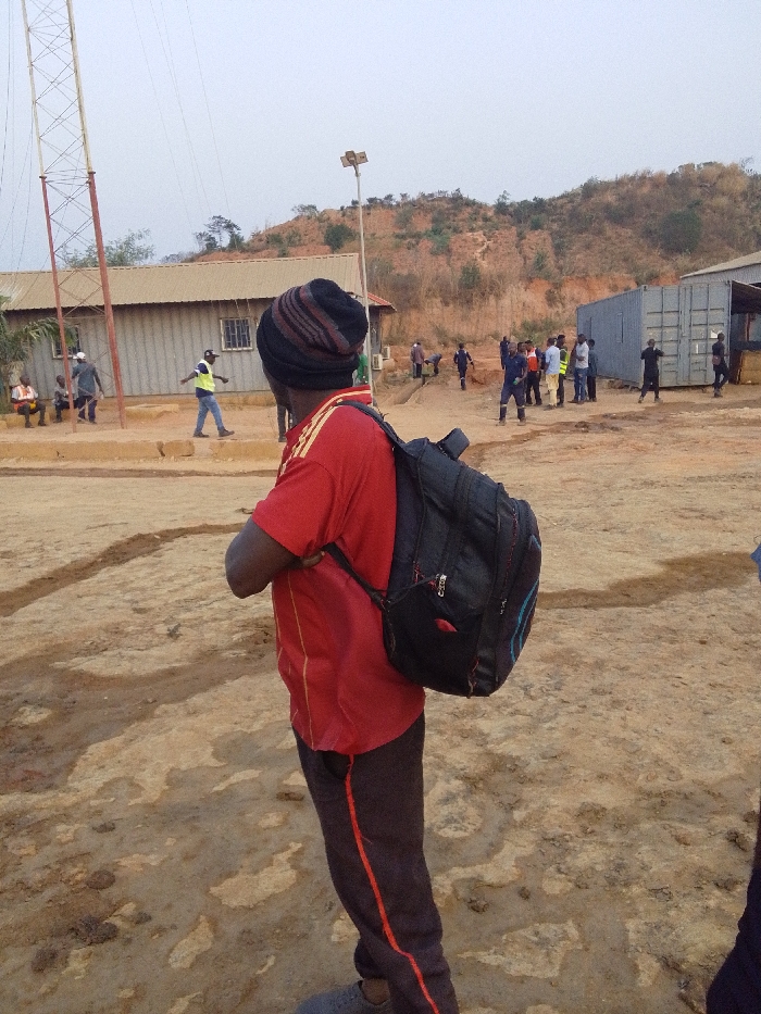 The image depicts an outdoor scene in Hoche, Nigeria, characterized by an unpaved, reddish-brown dirt ground with visible depressions and uneven patches. In the immediate foreground, facing away from the camera and slightly to the right, is a person wearing a red short-sleeved shirt with yellow shoulder accents, dark trousers with a red stripe, and a striped knit beanie (black, brown, red). This individual carries a black backpack with blue accents.
The midground features various structures and groups of people under an overcast sky. To the left, a tall metal lattice tower stands beside a single-story building with a light-colored corrugated metal roof and walls, equipped with barred windows. Several individuals are present near this building; one person in a light safety vest walks from left to right, while others stand or sit. Centered in the midground, a wooden pole topped with a solar panel and light fixture is visible. Further to the right, a cluster of approximately ten individuals, some appearing to wear work attire or safety vests, stand and interact. Behind them, multiple grey shipping containers are utilized as structures, with one appearing partially open. Another structure with a corrugated metal roof is partially visible on the far right.
The background consists of a prominent reddish-brown hill with sparse, dry vegetation, suggesting a semi-arid or excavated landscape. The overall environment appears to be an active work site or a transitional area.