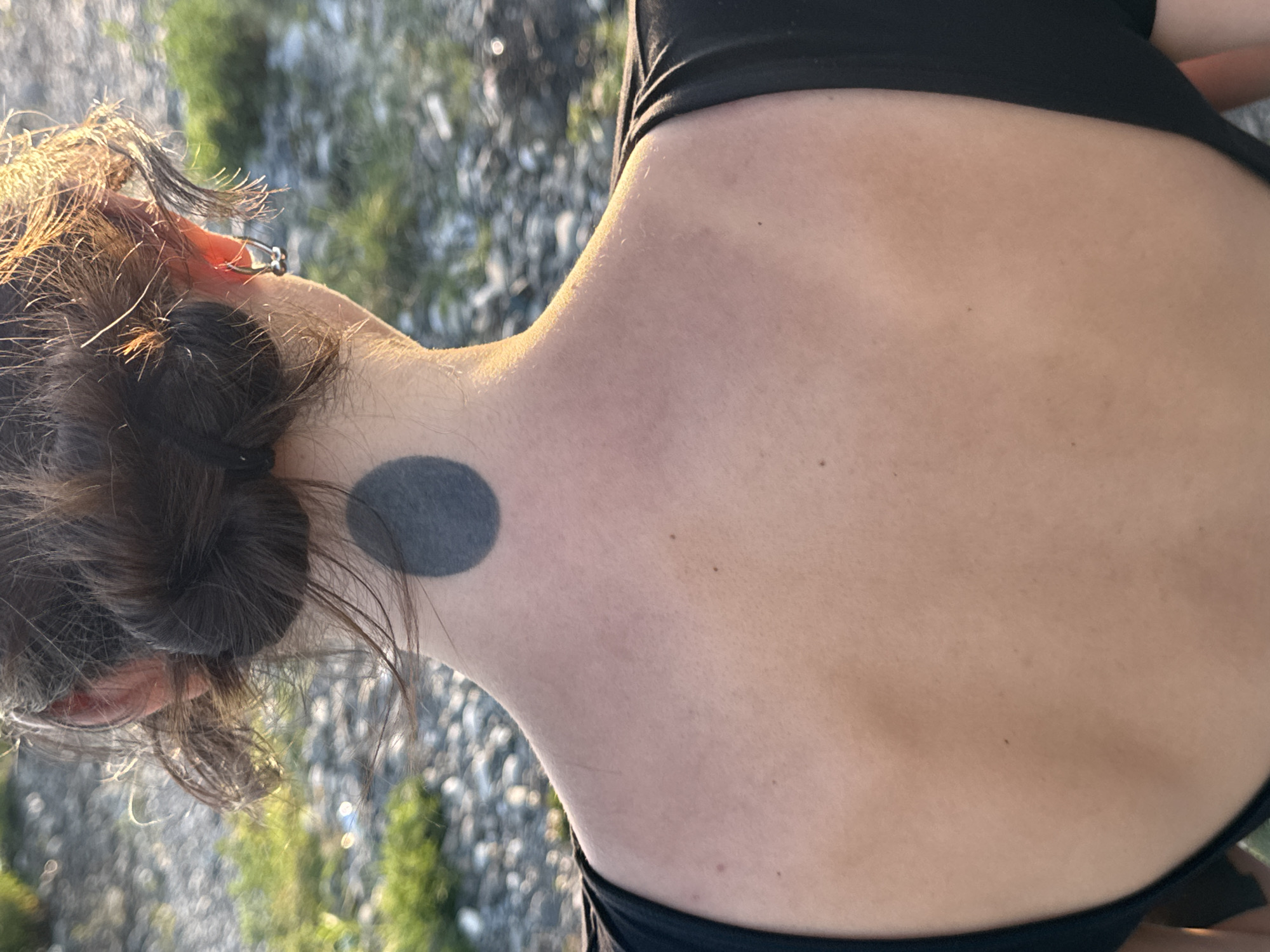The image shows the back of a person's head and neck, with their hair tied up in two buns. The person is wearing a black top. There is a black circular tattoo on the back of their neck. The background appears to be an outdoor setting with gravel and some greenery, possibly a park or a beach. The lighting suggests it is daytime. The image is cropped to focus on the person's upper back and head. No specific location within the city of Sochi, Russia, can be determined from the image.