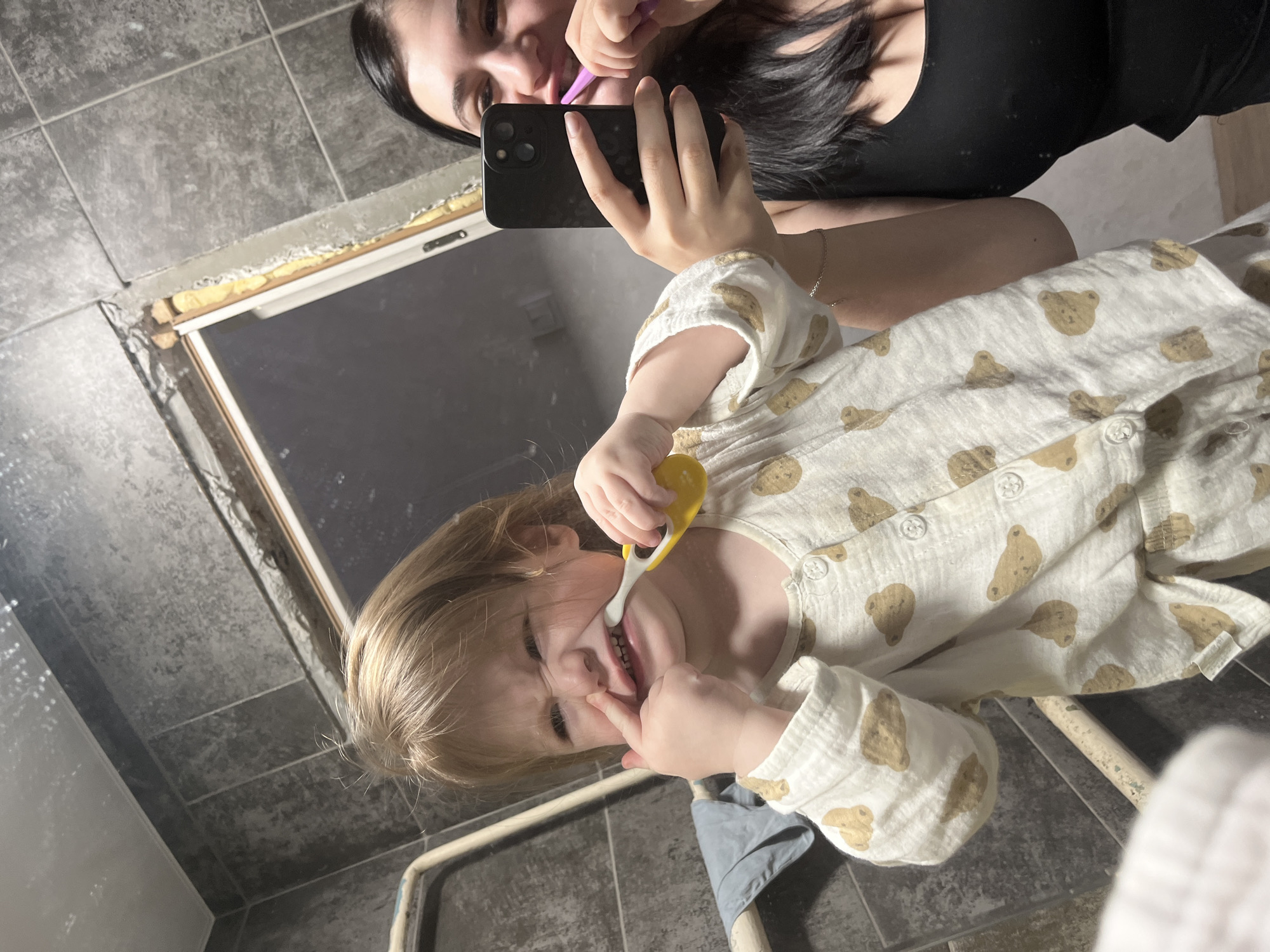 The scene depicts a mirror selfie taken in a tiled room, likely a bathroom, located in Chaikovsky, Russia. A young child with light-colored hair, wearing white pajamas patterned with brown bears, occupies the foreground. The child is holding a yellow toothbrush in their right hand, brushing their teeth, while their left index finger presses against their nose, distorting their upper lip and creating a crinkled facial expression. Reflected in the mirror behind the child, an adult with dark hair, wearing a black top, holds a dark-colored smartphone (equipped with three rear cameras) in their left hand to capture the image. The adult's right hand holds a purple toothbrush to her mouth. The background consists of dark grey, possibly stone-textured, wall tiles. To the immediate left, a light-colored rail or radiator is visible, with a blue-grey cloth draped over it. In the upper-right portion of the mirror's reflection, an unfinished window or doorway is apparent, showing exposed wooden framing and yellow construction foam. A folded white towel is partially visible in the bottom-left foreground.
