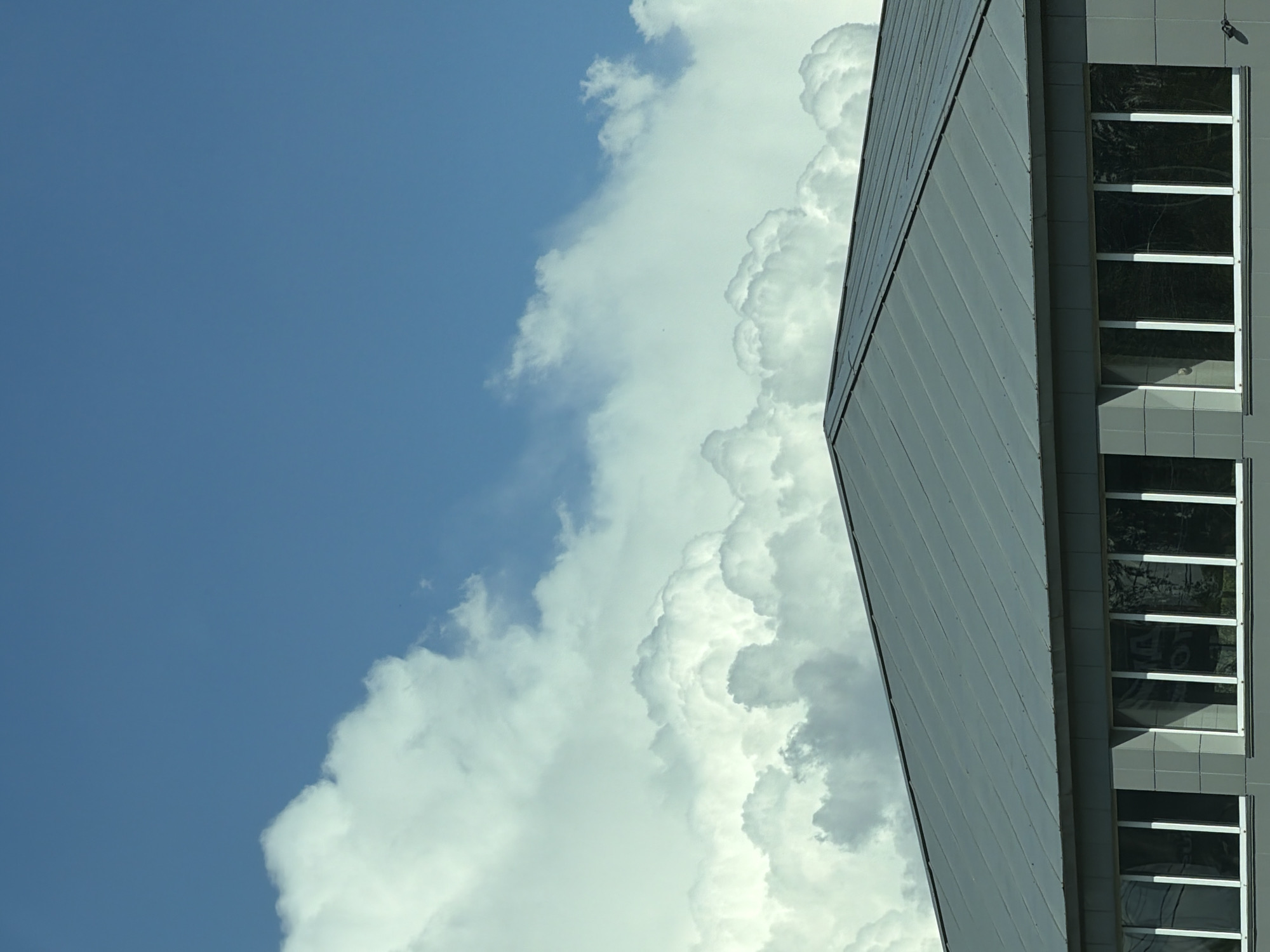 The image shows a building under a blue sky with voluminous cumulus clouds. The building's exterior appears to be made of gray panels and features vertical rows of windows with white frames. Reflections of trees or other foliage are visible in the windows. The exact location cannot be verified based solely on the image and without additional context or identifying information about Vereya, Russia.