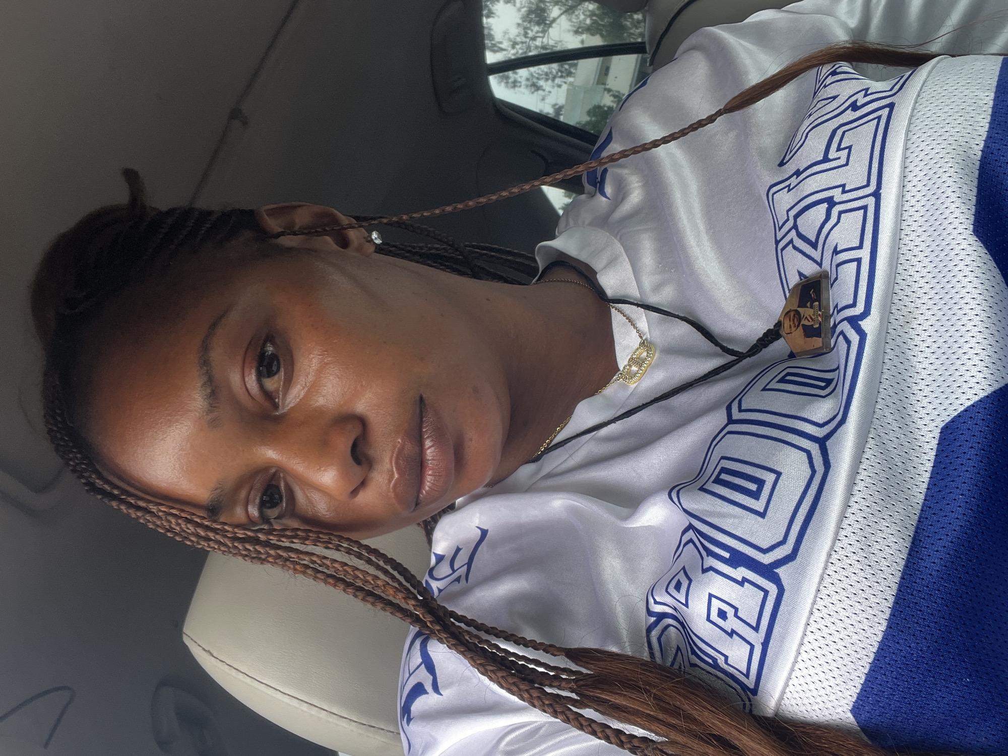 A close-up, medium-shot, eye-level selfie of a young Black woman with braided hair is taken from inside a vehicle. She is wearing a white jersey with blue trim and graphics. The woman is looking towards the camera with a neutral expression. She has a gold necklace with a pendant and a smaller pendant on a black string. A small, clear earring is visible in her left ear. The background of the image shows the interior of a car, including the headrest of a car seat, and out the window to a blurry background that suggests an outdoor, urban environment. There is no specific identifiable landmark to confirm the location in Jos, Nigeria.