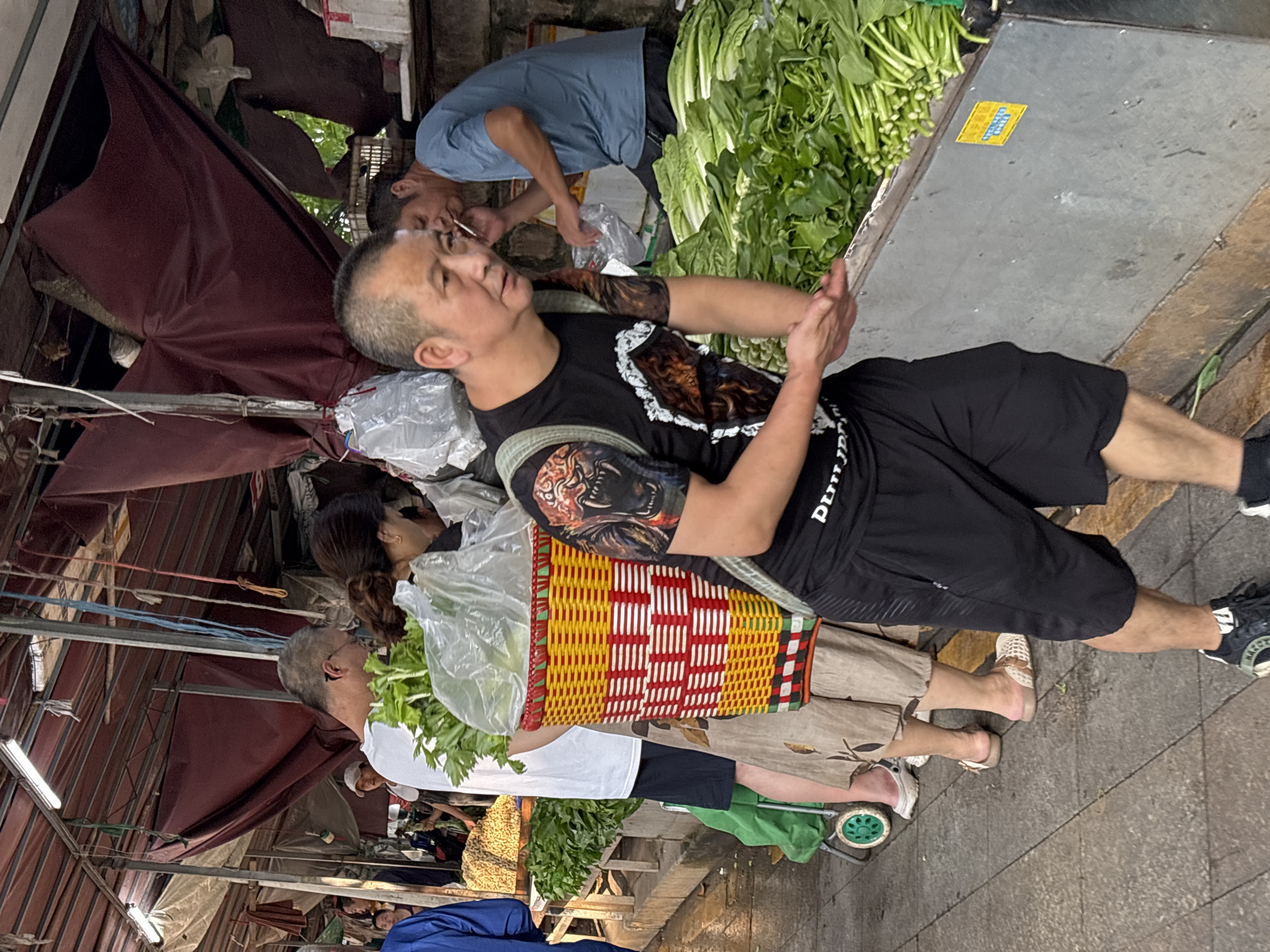 The image depicts an outdoor market scene in Jiangjin, China. In the foreground, a man with a tattoo on his arm is walking towards the right, carrying a backpack and wearing a black t-shirt and shorts. He appears to be reaching out with his left hand. Behind him, a woman in a patterned skirt carries a large yellow and red woven basket filled with green vegetables. To the left, a person in a white t-shirt and dark shorts stands with a cart. Further in the background, another man in a blue t-shirt is speaking on a cell phone and smoking. The market stalls are covered with tarps, and various fresh produce, including leafy green vegetables, are displayed on a metal counter on the right. The ground is paved with cobblestones.