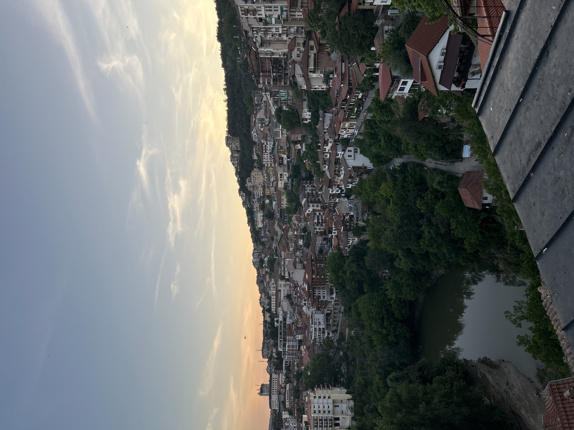 This is an aerial, wide-angle photograph of Veliko Tarnovo, Bulgaria, likely taken at dusk. The image is oriented vertically, with the sky occupying the left half and the town and surrounding landscape on the right.
The sky is a gradient of pale blue to soft yellow near the horizon, with wispy clouds scattered across it. Several small, dark specks are visible in the sky, possibly birds or distant aircraft.
The town is built on a steep hillside that slopes down towards a body of water, likely the Yantra River. The buildings are densely packed, with a mix of traditional terracotta-tiled roofs and more modern structures. Many of the buildings are white or light-colored, with visible windows and balconies. The architecture suggests a historic European town.
Dense green trees surround the town and the water, creating a lush, natural setting. The water's surface is dark and reflective, mirroring the surrounding foliage and sky. In the foreground, a portion of a dark, textured roof is visible, suggesting the photo was taken from a high vantage point, possibly a rooftop or a balcony.
The overall impression is one of a picturesque town nestled within a natural landscape, bathed in the soft light of twilight. There are no visible people or overt activities, but the scene evokes a sense of peacefulness and scenic beauty. No text is visible in the image.