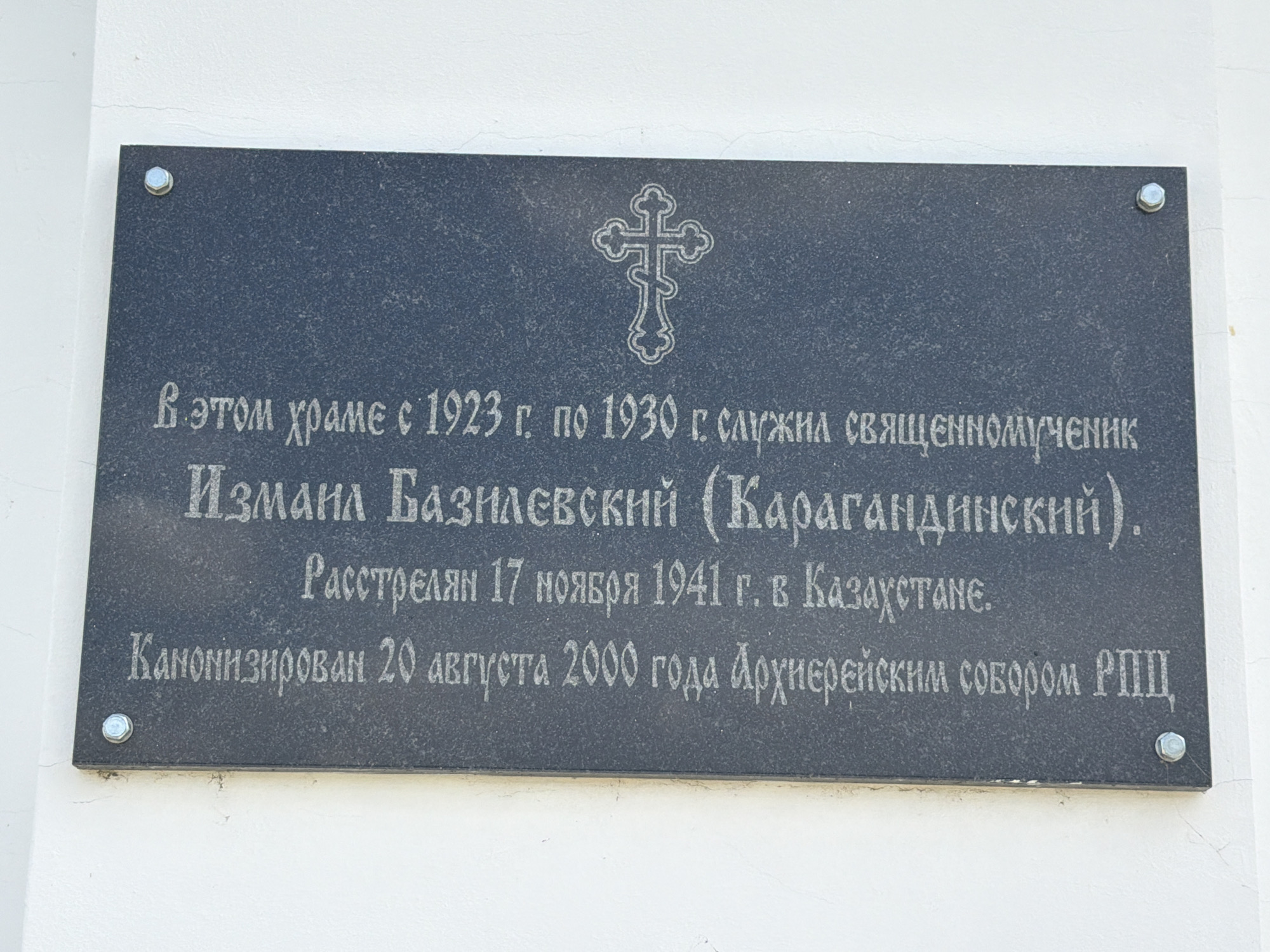 A dark granite plaque is affixed to a white stucco wall with four bolts. At the top center of the plaque, an ornate cross is etched. Below the cross, text in white lettering reads: "В этом храме с 1923 г. по 1930 г. служил священномученик Измаил Базилевский (Карагандинский). Расстрелян 17 ноября 1941 г. в Казахстане. Канонизирован 20 августа 2000 года Архиерейским собором РПЦ." The plaque is located on the Church of the Transfiguration in Skorniákovo, Russia.