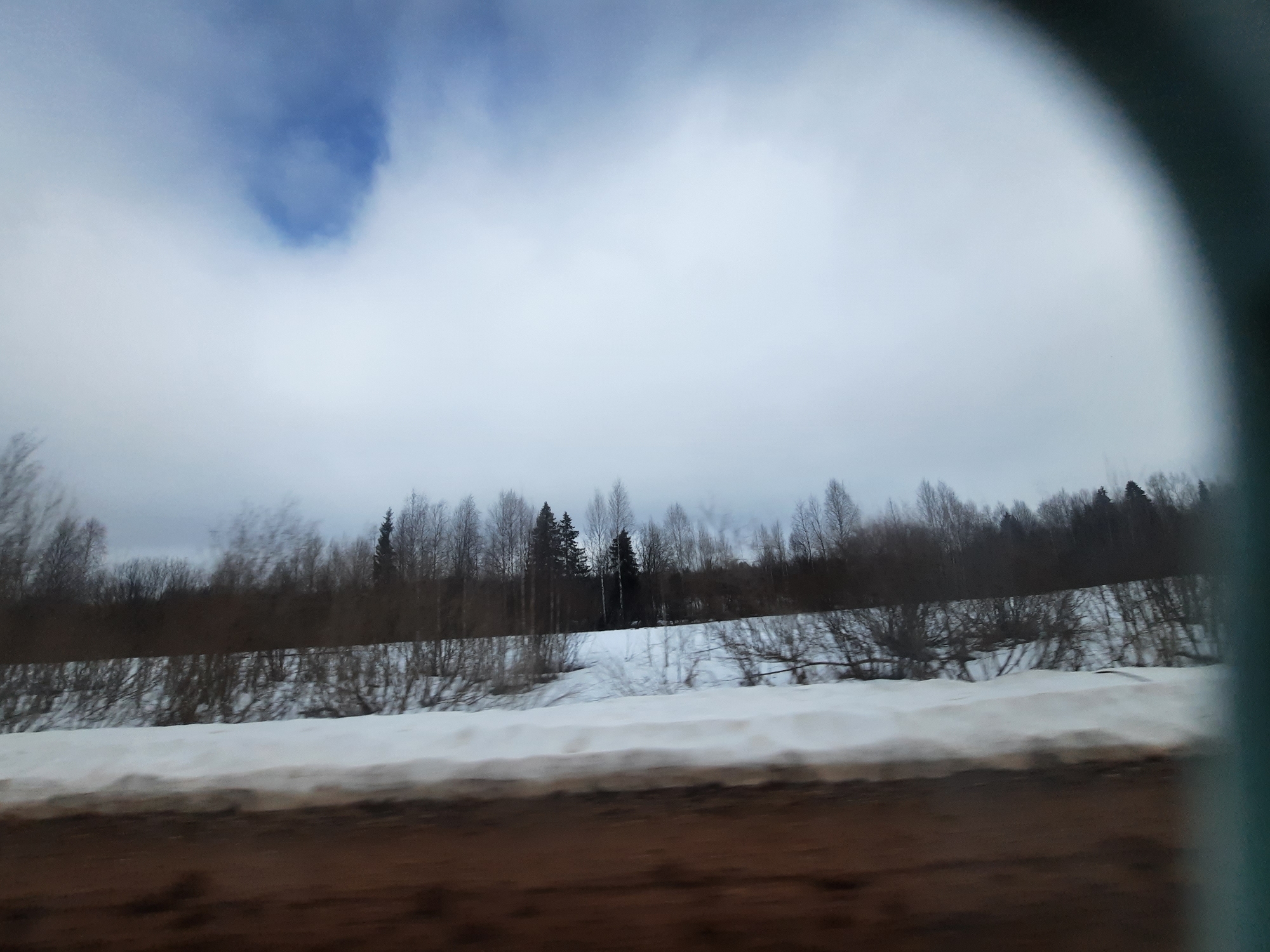 The image shows a winter scene in what appears to be a rural or suburban area near Архангельское, Russia. In the foreground, there is a blurry section of a road or ground, suggesting the photo was taken from inside a moving vehicle. A snowdrift is visible between the road and a line of trees and bushes. The trees are mostly bare, indicating winter. The sky is overcast with clouds, with a small patch of blue visible.