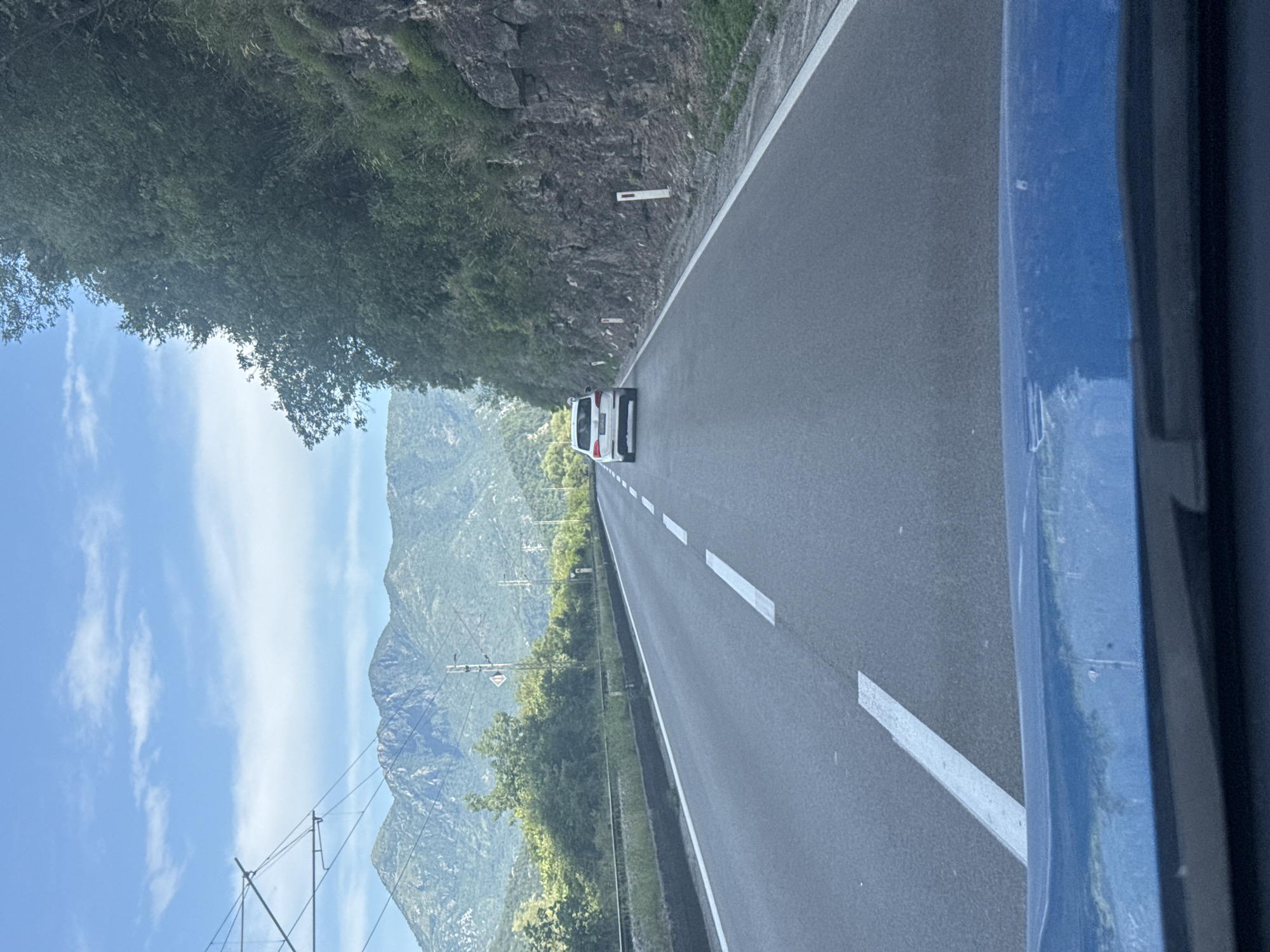 The image captures a view from a vehicle traveling on a paved road that runs alongside a steep, rocky mountainside covered in dense green foliage. A white van is visible ahead on the road. The road itself is marked with white dashed lines. To the left of the road, the terrain slopes upwards towards the mountain. In the distance, more mountains can be seen under a bright blue sky with scattered white clouds. Power lines with insulators are also present in the landscape. The image is oriented such that the road and the scene appear to be running vertically. The lighting suggests it is daytime, likely late morning or afternoon, with clear weather. The overall impression is one of travel through a scenic, mountainous region.