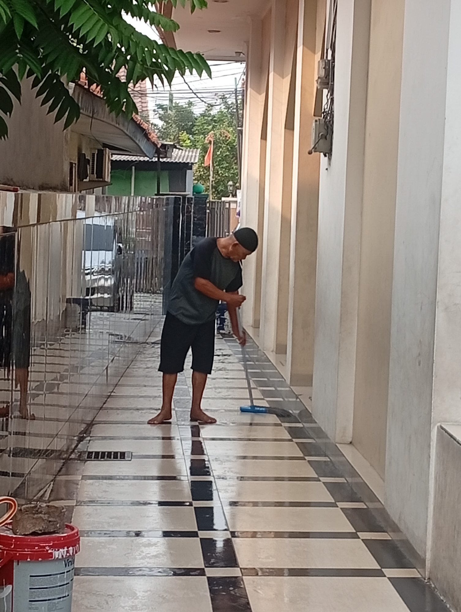 An individual is mopping a tiled walkway in front of a building in Bekasi, Indonesia. The person, wearing a dark t-shirt and shorts, is bent over, actively sweeping the floor with a blue-handled mop. The walkway features a checkerboard pattern of light and dark tiles, reflecting the sunlight. To the left of the walkway, there is a mirrored wall, creating a distorted reflection of the surroundings. A red bucket with white paint and some rocks is visible in the lower left corner of the image. The building's exterior consists of light-colored pillars and walls, with visible electrical conduits and meters attached to the wall. Lush green foliage hangs from above on the left side of the image, casting shadows on the walkway.