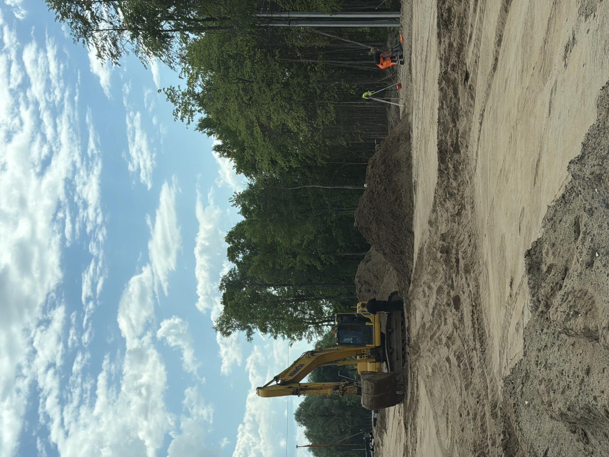 This image depicts a construction site in Kabanikha, Russia, on a partly cloudy day. A yellow Komatsu excavator is parked on sandy ground, with its bucket lowered. A construction worker in dark clothing stands beside the excavator. In the background, a large pile of dirt is visible, along with a line of trees. Further to the right, another construction worker in an orange safety vest and pants is operating surveying equipment. A tripod with a green prism attachment stands next to him. Electrical poles are also present on the right side of the image. The ground is uneven, with tire tracks from heavy machinery evident in the sand.
