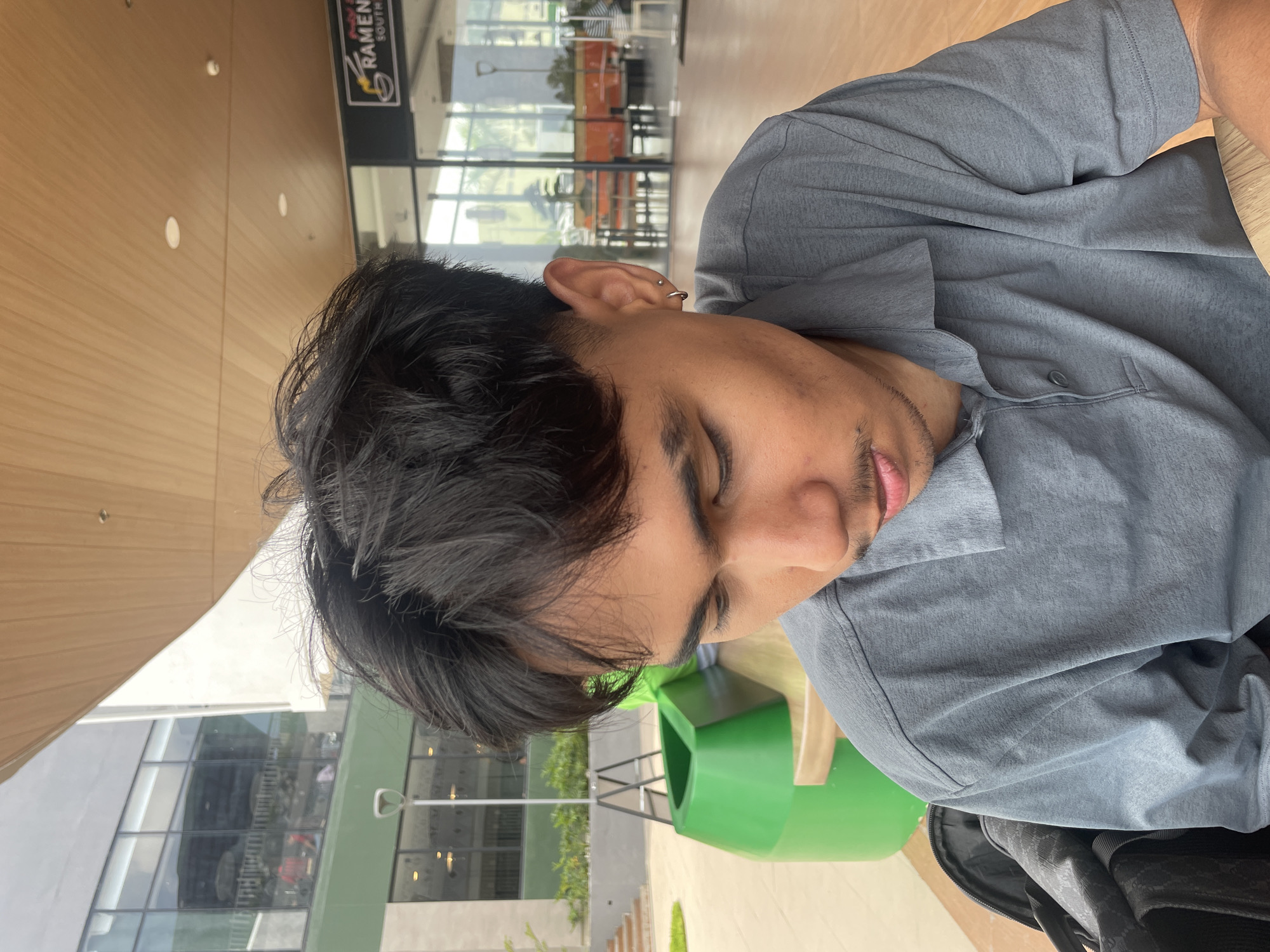A close-up, eye-level shot focuses on a young Filipino man with dark, messy hair and a light mustache, looking down. He is wearing a grey polo shirt. He has a small hoop earring in his left ear. He appears to be sitting at an outdoor table, with a black bag on his left. In the background, there is a bright green trash receptacle and a building with large glass windows. A sign for "RAMEN SOUTH" is visible on the building. The overall setting suggests an outdoor dining or leisure area within a commercial establishment in Davao City, Philippines. The lighting is bright, indicating it is daytime.