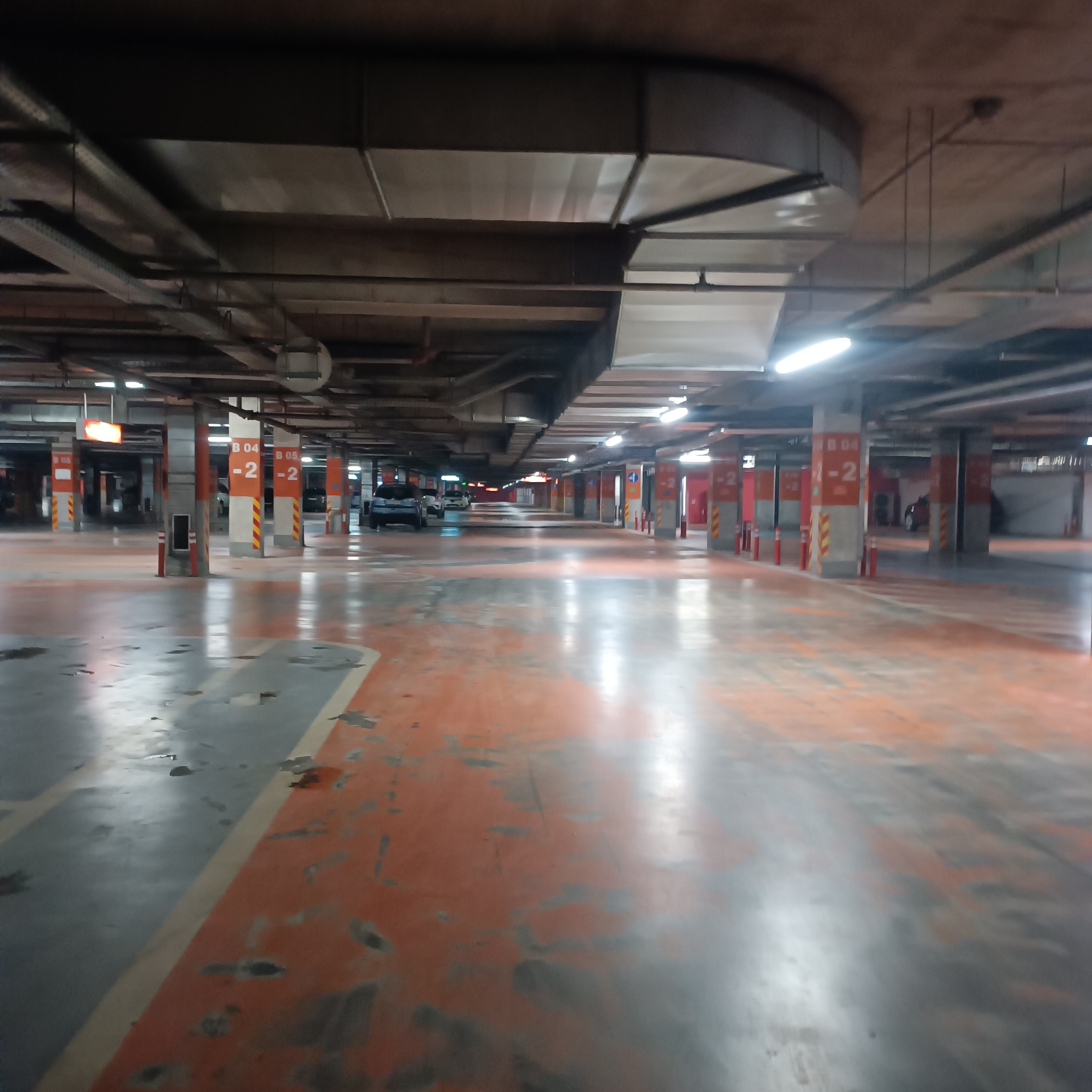 This image depicts an underground parking garage, likely located in Sofia, Bulgaria, characterized by its utilitarian design and extensive infrastructure. The scene is dominated by rows of concrete pillars supporting a low ceiling, with the floor painted in alternating sections of orange and gray, showing visible wear, peeling paint, and damp or stained patches.
Overhead, a complex network of large rectangular air ducts, various pipes, and electrical conduits runs across the ceiling. Numerous bright fluorescent light fixtures illuminate the space, creating strong reflections on the polished or wet sections of the floor.
Several cars are parked further down the aisles, indicating the facility is in use, though no people are visible, suggesting a quiet or off-peak moment. The concrete pillars are marked with signs, clearly displaying "B 04 -2" and "B 05 -2," which designate specific parking sections and indicate that the image was taken on the second basement level. Some pillars are also adorned with yellow and red striped markings, possibly for visibility or safety. Traffic cones are placed intermittently beside the pillars. The overall impression is one of a vast, functional, and well-lit, albeit somewhat aged, underground parking facility.