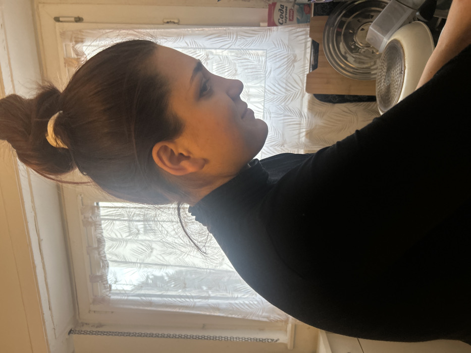 The image captures a close-up profile view of a person, likely female, facing towards the right side of the frame. Her dark brown hair is neatly pulled back into a high bun at the back of her head, secured with a light-colored (possibly off-white or cream) crescent-shaped hair clip or barrette. She has a smooth forehead and a visible ear, and her facial expression appears calm and contemplative as she looks slightly upwards.
She is wearing a dark, possibly black, long-sleeved top with a distinctive high, ruffled neckline, giving it a turtleneck or mock-neck style.
The background primarily features a large window with sheer white curtains adorned with a repeating pattern of light-colored leaves or ferns. Bright light streams through the window, indicating it is daytime. Outside the window, a blurry view suggests either outdoor scenery or simply bright light.
To the right side of the person and behind her, various kitchen items are visible. These include a shiny silver, metallic object, which appears to be the lid of a pot or pressure cooker, reflecting light. Below it is a wooden cutting board, and further to the right, a white pan or dish with a textured or perforated bottom is partially seen. A box or container with Cyrillic text, clearly showing "СОДА" (SODA), is also visible on a shelf or surface in the upper right portion of the background.
The setting appears to be an indoor kitchen or dining area, illuminated by natural light from the window. The person's posture suggests she might be engaged in an activity, possibly observing something in front of her or simply standing in the kitchen environment.