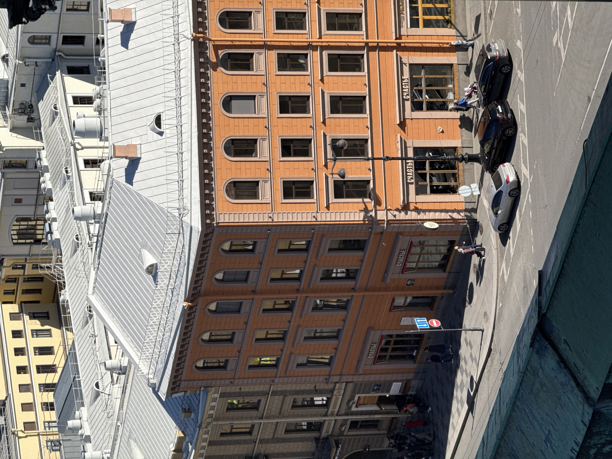 This photograph is taken from an elevated perspective, seemingly from atop a building. It captures a street corner in what appears to be Saint Petersburg, Russia. Buildings with ornate, symmetrical facades line the street, featuring multiple windows and a color palette that includes shades of orange and yellow. A building on the right has signs in Russian script.
The street shows pedestrian and vehicular traffic. There are several cars parked alongside the building. The presence of pedestrian crossing signs suggests an urban environment, indicating that people may be crossing the road.
The photograph captures a slice of life in a Russian city, with architectural details that signify the area's historical significance. The weather appears to be clear and sunny, casting shadows that define the forms and shapes in the scene.