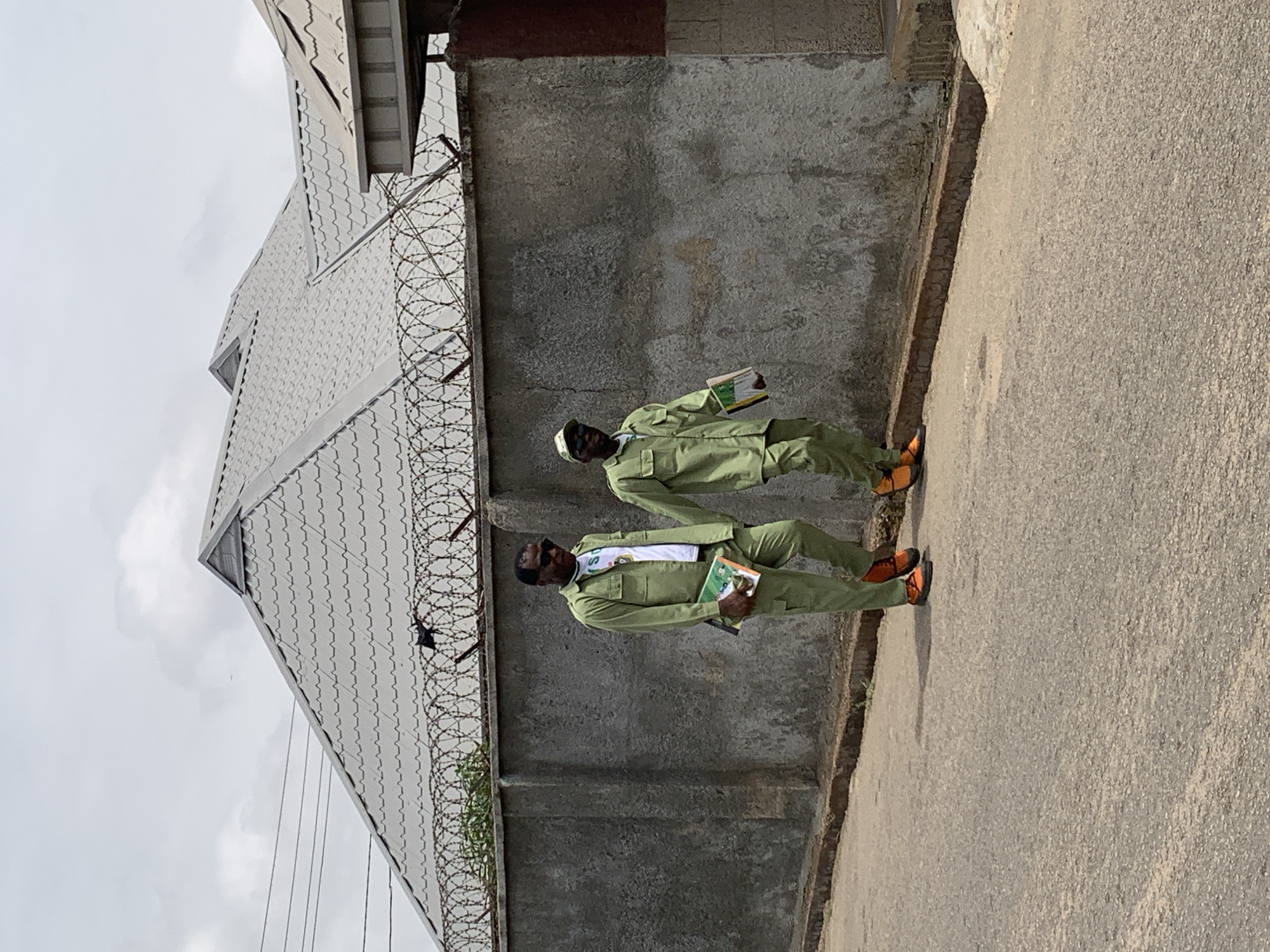Two people in green uniforms and orange boots walk along a concrete sidewalk, adjacent to a grey concrete wall topped with barbed wire. A grey roof is visible behind the wall. The location is in Jos, Nigeria. The individuals are holding books.