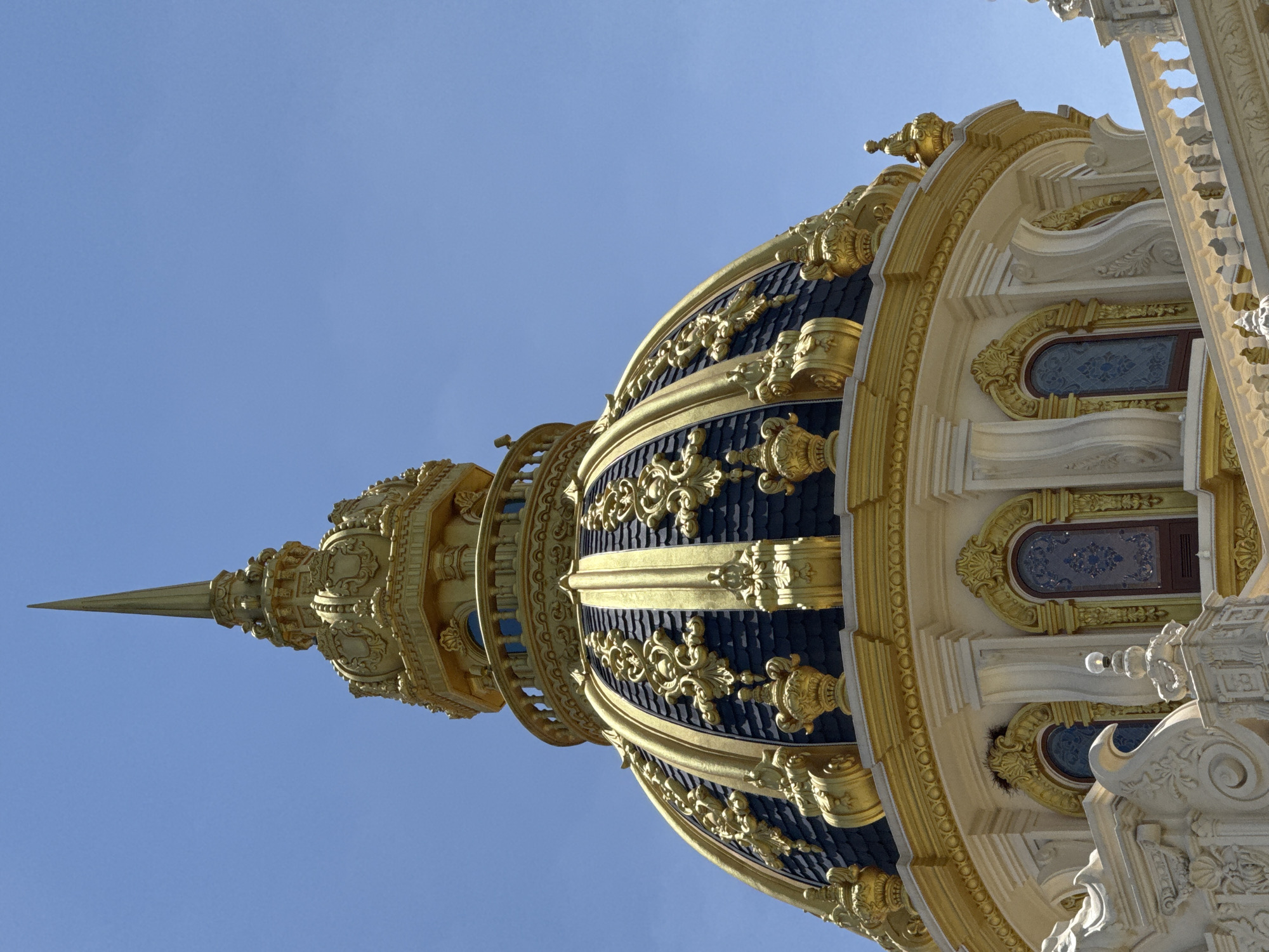 This is a close-up, low-angle shot of the golden dome of the Dinh II (Villa Dinh II), a French colonial villa located in Da Lat, Vietnam. The dome is ornate, featuring dark blue shingle-like roofing with intricate gold detailing, including floral motifs and finials. The golden structure extends upwards to a sharp spire. Below the dome, the building's facade is visible, adorned with white stucco decorations, arched windows with decorative glass, and a balustrade. The sky is clear and blue, indicating daylight. There are no people visible in the image.