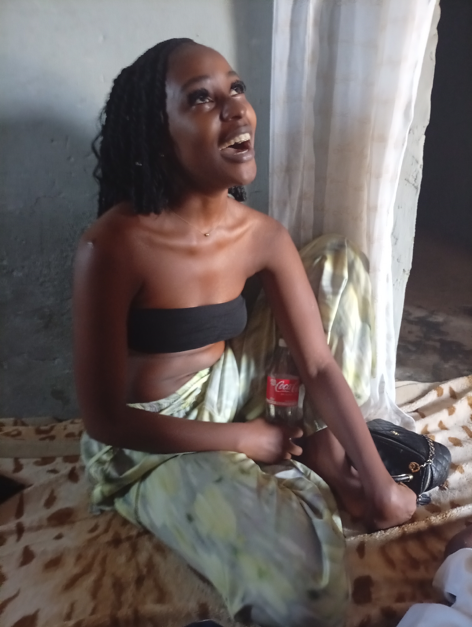 A young woman is pictured indoors, seated on a patterned blanket or rug, looking upwards with an open mouth, indicative of laughter or a joyful expression. She has dark skin and her hair is styled in dark braids. She wears a black bandeau top, a delicate gold pendant necklace, and a light-colored, loosely patterned fabric wrapped around her waist, resembling a skirt or wrapper. In her right hand, she holds a glass Coca-Cola bottle, prominently displaying the red and white "Coca-Cola" logo.
Behind her, a textured gray wall is visible, and to her right, a sheer white curtain hangs, partially revealing a dark opening, possibly a doorway or window. A black quilted handbag with a gold chain strap rests on the patterned surface beside her. The lighting is soft and even, suggesting a casual, relaxed indoor environment. The scene appears to capture a moment of genuine amusement or happiness. The location context places this scene in Sabon Tasha, Nigeria.