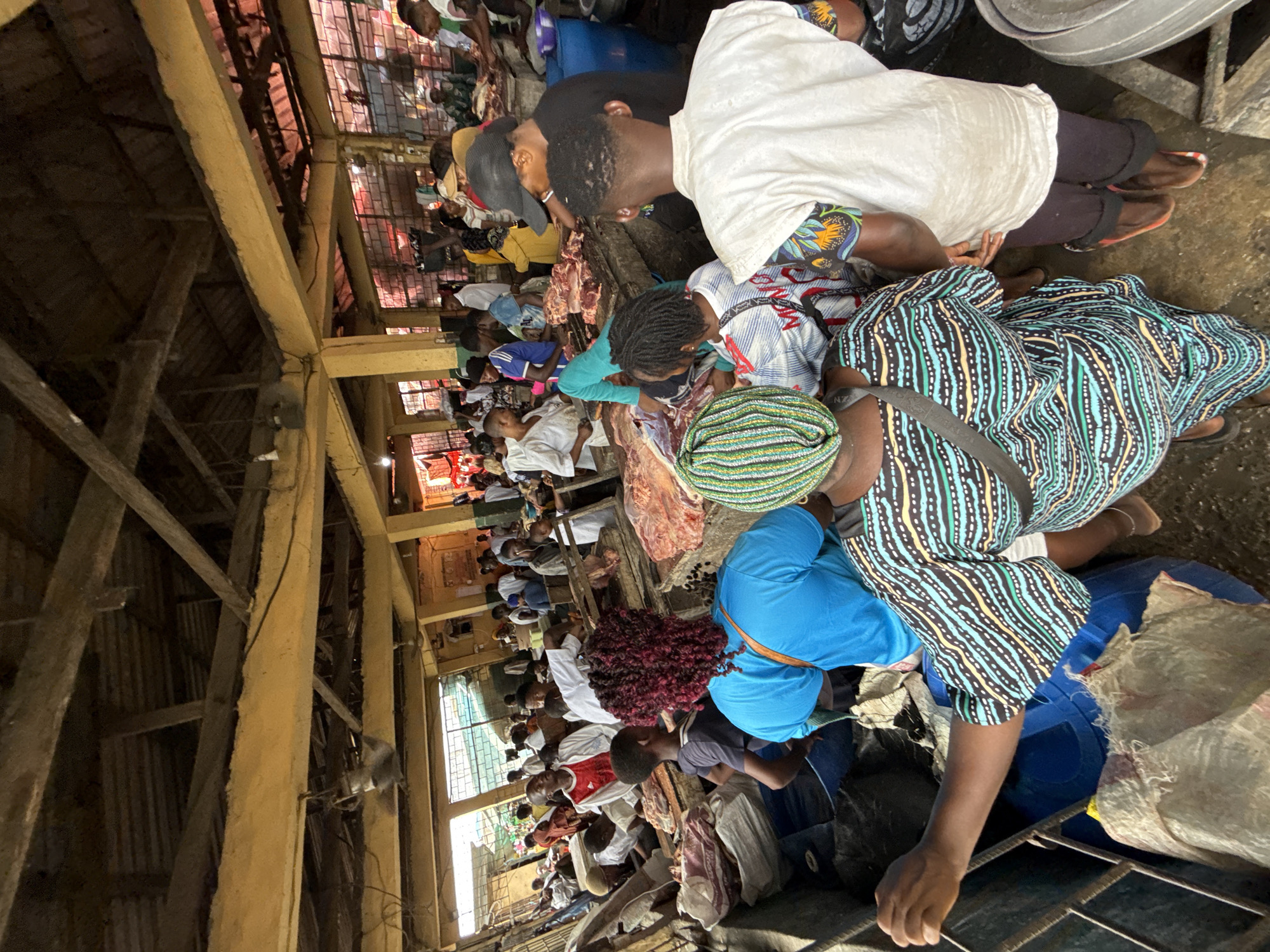 The image displays a bustling indoor market scene within Lekki, Nigeria, characterized as a meat market. The structure features a corrugated metal roof supported by exposed wooden beams and yellow-painted concrete pillars and crossbeams. Natural light enters from openings in the background, supplemented by overhead incandescent bulbs.
Numerous individuals, consisting of men and women, populate the dense space. In the immediate foreground, a person wearing a multi-patterned green, black, and blue garment and a matching headwrap is visible from the back, holding onto a metal rail. To their right, another individual, also seen from the back, wears a white short-sleeved top with colorful patterned sleeves over dark pants and open-toed footwear. A third person, facing away, has red curly hair and wears a bright blue short-sleeved top. Blue plastic barrels and white sacks are visible at ground level near these individuals.
The midground and background reveal multiple market stalls where large quantities of raw, reddish-pink meat are displayed on elevated wooden or concrete counters. Various people are congregated around these displays. Some individuals, presumably vendors, are actively engaged with the meat, while others appear to be customers observing or interacting. Attire varies, including plain white shirts, t-shirts, and head coverings. One person in a black cap and black t-shirt is positioned behind a meat display. The floor surface appears to be concrete.