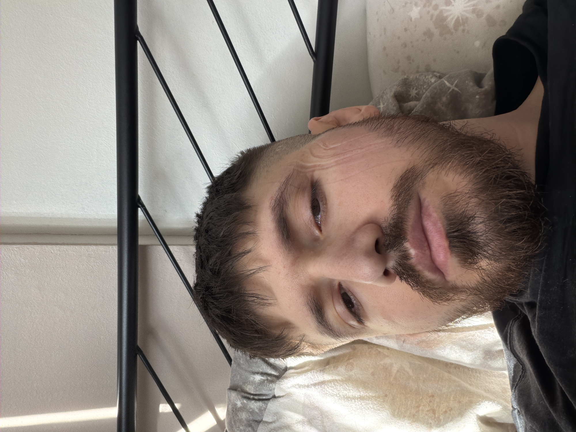 A close-up, slightly skewed portrait shows a light-skinned man with dark hair and a full, short beard. He appears to be lying on a bed with a patterned comforter and a portion of a black metal bed frame is visible in the upper left. He is wearing a dark-colored shirt. No elements in the image suggest it was taken in Pentre, United Kingdom.