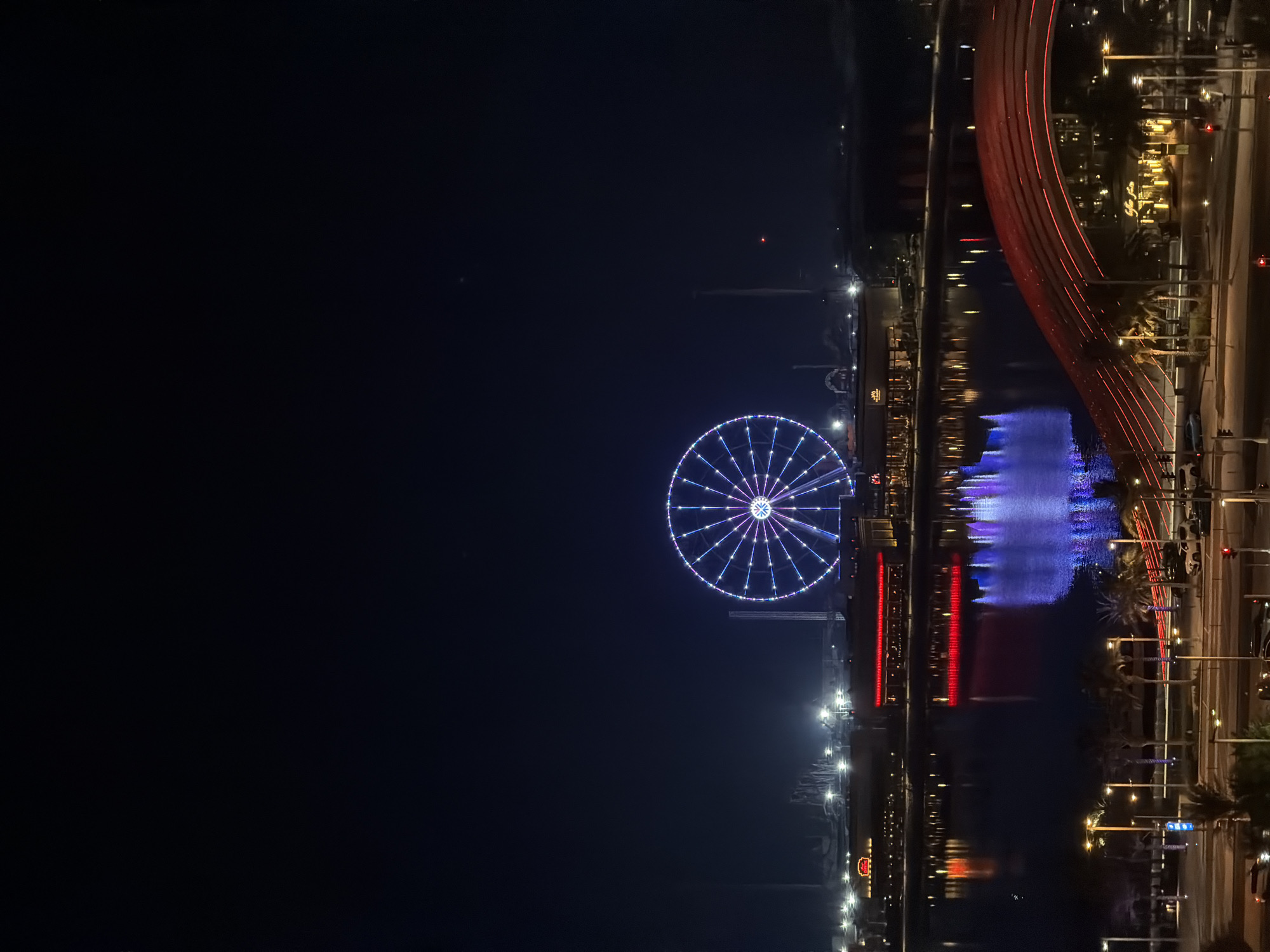 An eye-level, medium shot captures a Ferris wheel at night in an unknown city. The Ferris wheel, illuminated with blue and white lights, is the central focus of the image. The wheel is positioned above a body of water that reflects the lights of the surrounding buildings and the Ferris wheel itself. In the foreground, a red, arch-shaped bridge with red lights spans across the scene, with palm trees lining the area. Buildings with red and yellow lights are visible on the waterfront. The sky is dark, indicating nighttime. The scene is devoid of people.
