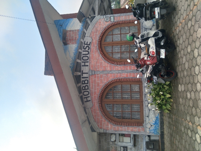 The image displays the exterior of a building identified as "HOBBIT HOUSE" located in Sukatani, Indonesia. The structure features a main facade painted to mimic red brick, with a gray faux stone base. Two large arched windows, fitted with brown wooden frames and interior curtains, are central to the facade. Above these windows, a white sign with dark lettering reads "HOBBIT HOUSE." A dark, sloped roof with a pronounced overhang covers the building, beneath which a mural depicting a blue sky and green landscape is painted on the upper wall. A blue stylized wave pattern is painted along the building's base. To the left of the main windows, a gray wall section includes a mounted white box and a small white sign marked "9.2." To the right, a circular white sign bearing the number "99" is affixed to the wall.
Three motor scooters are parked on the gray hexagonal paver driveway directly in front of the building. From left to right, these are: a dark red scooter (license plate B 3528 FZF), a white scooter with a green helmet resting on its seat (license plate B 5682 OYX), and a black scooter (license plate B 5572 ZBD). A cluster of green plants with white flowers is situated between the red and white scooters, positioned in front of the building's base. The scene is illuminated by natural light under a clear, light blue sky. No individuals are visible.