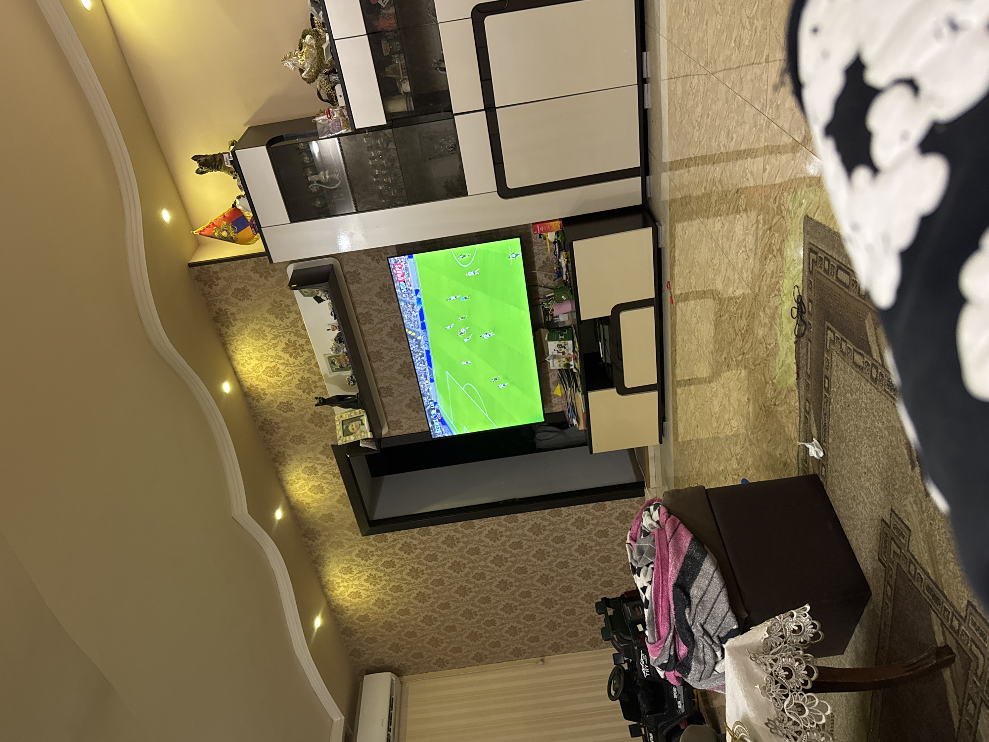 The image depicts an interior space, likely a living room, featuring a television displaying a soccer game. The room is furnished with a modern entertainment center, a rug, and an ottoman with draped textiles. Decorative elements include figurines and framed pictures. Wallpaper adorns the walls.
While the location is assumed to be Yerevan, Armenia based on potential contextual information, no specific details within the image confirm this claim.