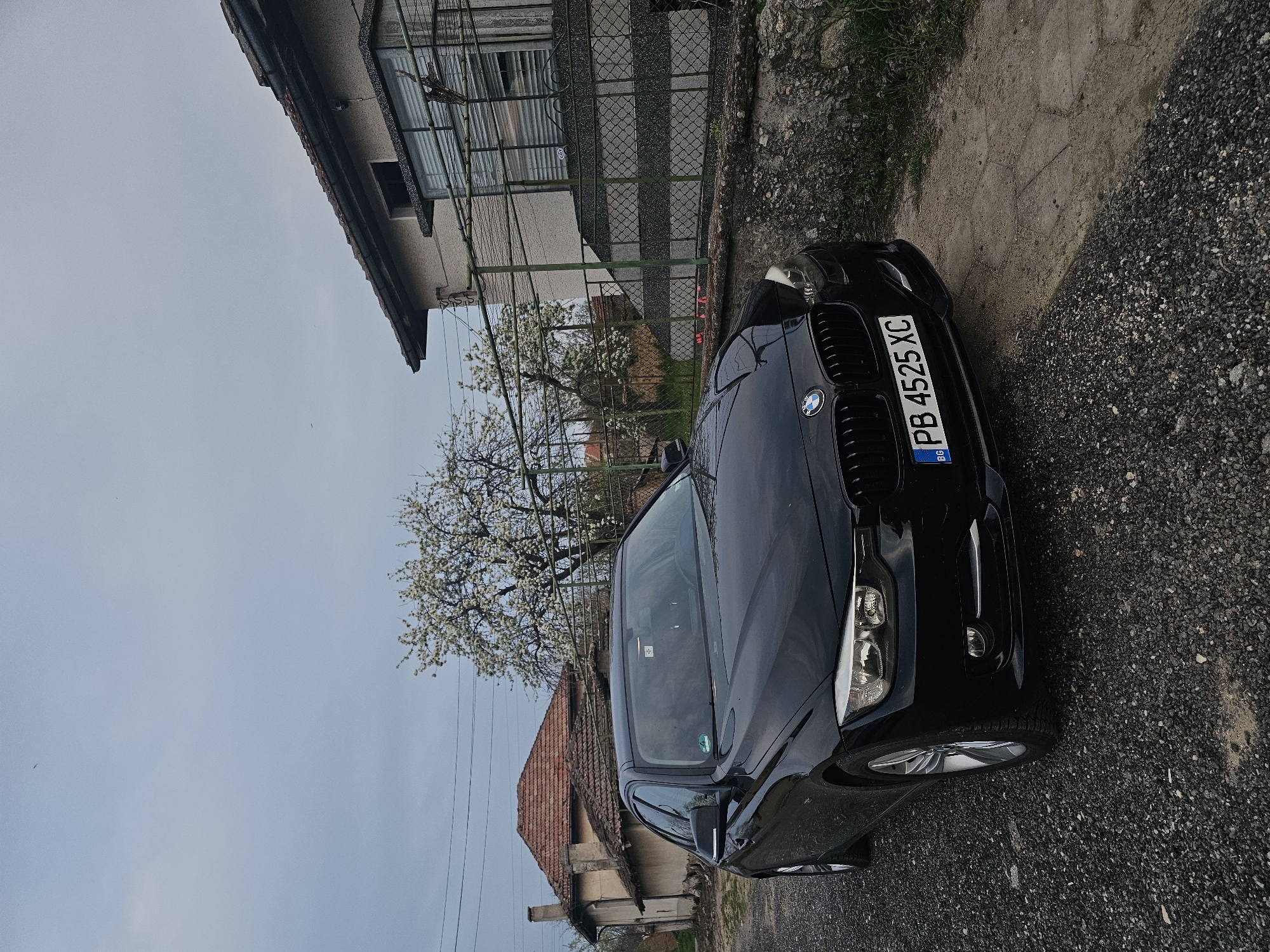 A black BMW passenger vehicle is parked on a dirt road with gravel, and is near a residence with an adjacent chain-link fence, and a tree is partially visible to the left of the vehicle, which has license plate PB 4525 XC, potentially in Krasnovo, Bulgaria.