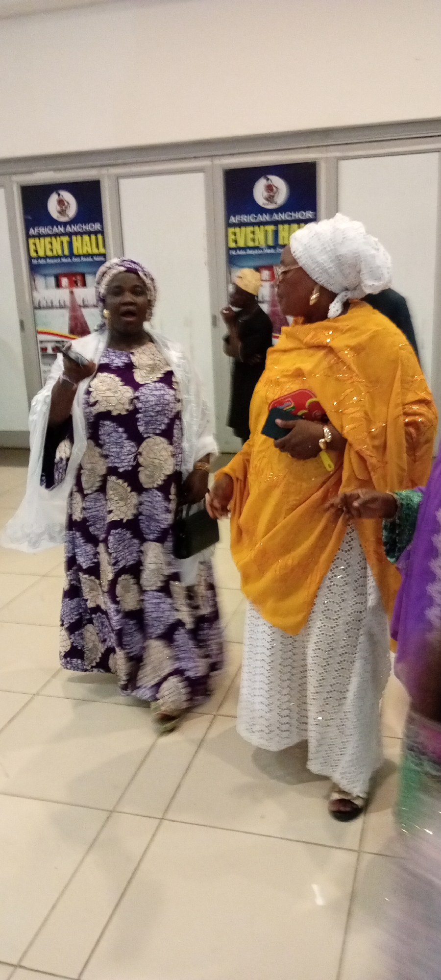 Two women are standing and facing forward. The woman on the left is wearing a purple and white patterned dress, a white shawl, and a purple and white headscarf. She is holding a phone in her left hand. The woman on the right is wearing an orange headscarf, an orange shawl with gold sequins, and a white lace skirt. She is holding a clutch purse in her right hand. Behind them, a sign reads "AFRICAN ANCHOR EVENT HALL." The floor is tiled with light beige squares. The scene appears to be an indoor event hall in Gandu Albasa, Nigeria.