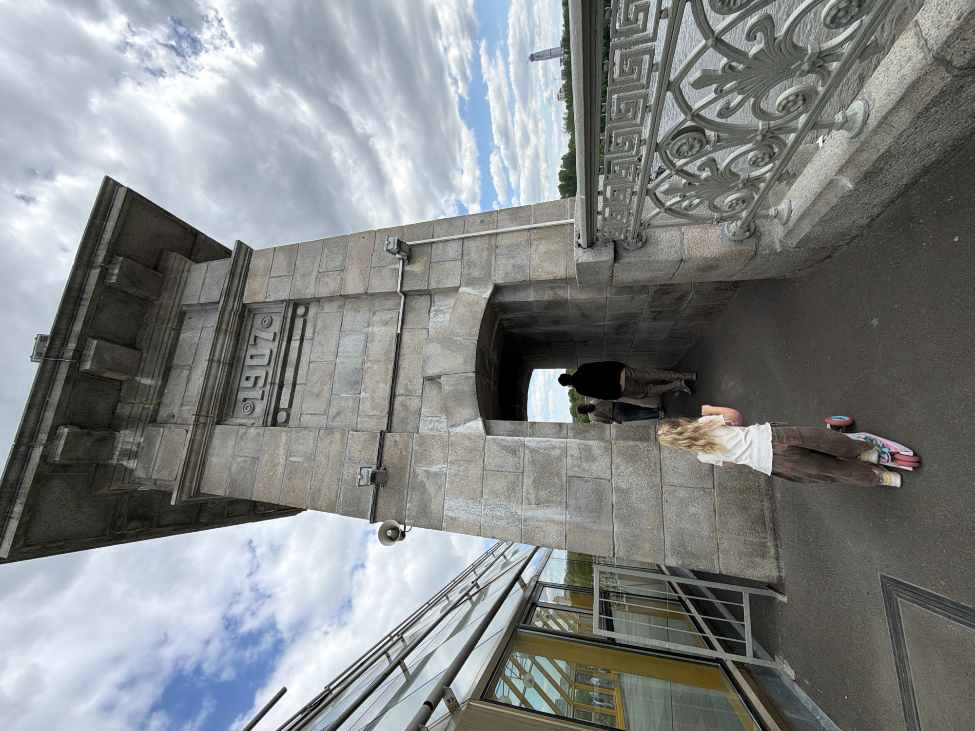 The image captures a scene from Moscow, Russia, depicting a stone archway structure with the number "1907" inscribed on its facade. Under the arch, two people are walking away from the camera's perspective. To the right of the archway, a young girl with blonde hair is seen lying on a scooter. The area is a walkway, possibly a bridge or overpass. A modern glass-walled building is visible in the lower portion of the frame. Above, the sky is filled with clouds.